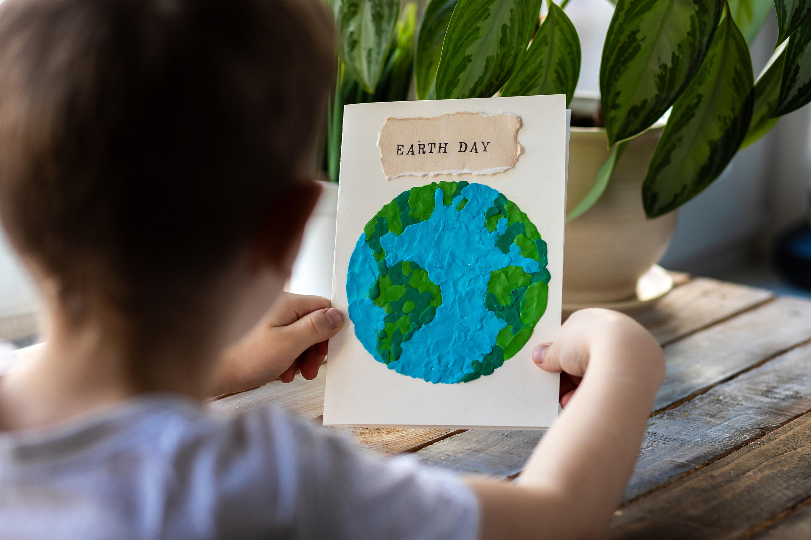 Ein Kind, das eine Karte mit der Erde und der Aufschrift "EARTH DAY" hält. Im Hintergrund sind grüne Pflanzen auf einem Holztisch zu sehen.