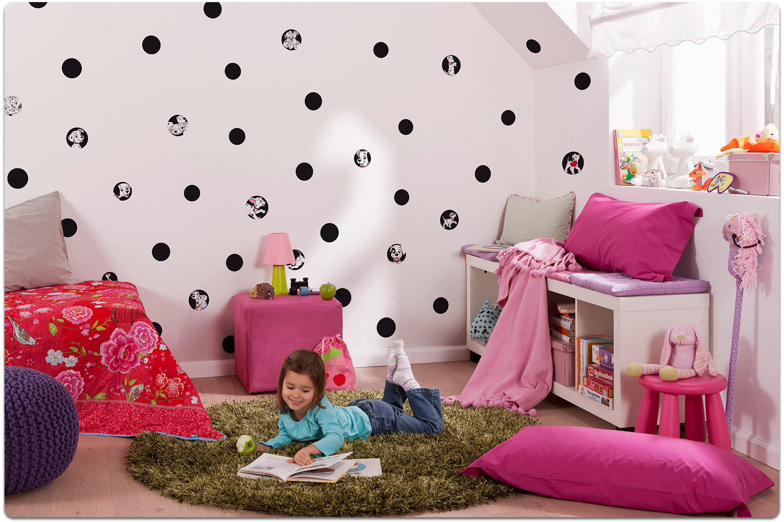 Kind liegt auf grünem Teppich in einem Kinderzimmer mit rosa und roten Kissen, einem Bücherregal und einer Wand mit schwarzen Punkten.