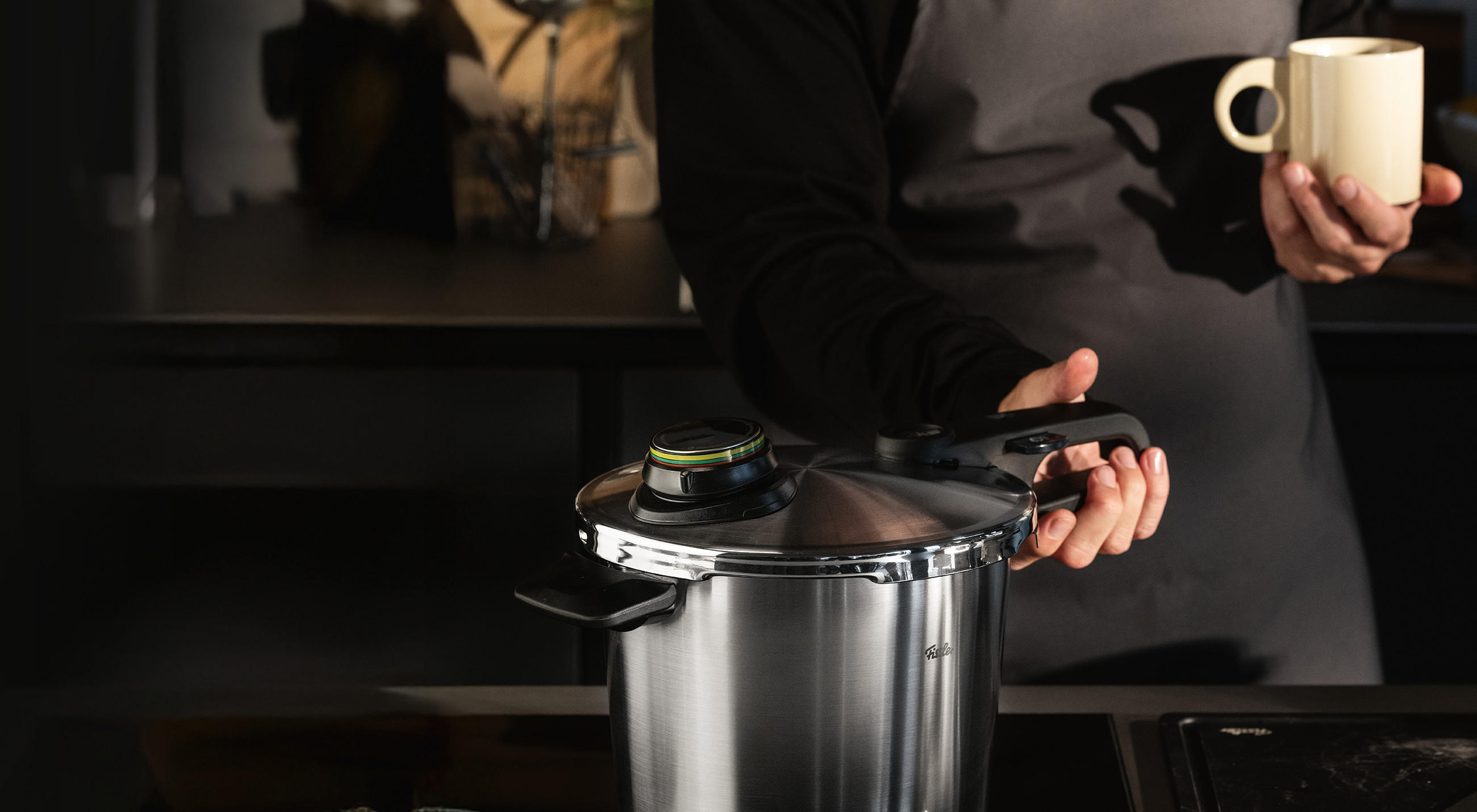 Person in dunkler Kleidung hält den Griff eines FISSLER Edelstahl‑Schnellkochtopfs auf dem Herd; am Deckel ist die farbige Druckanzeige sichtbar, in der anderen Hand eine helle Tasse.