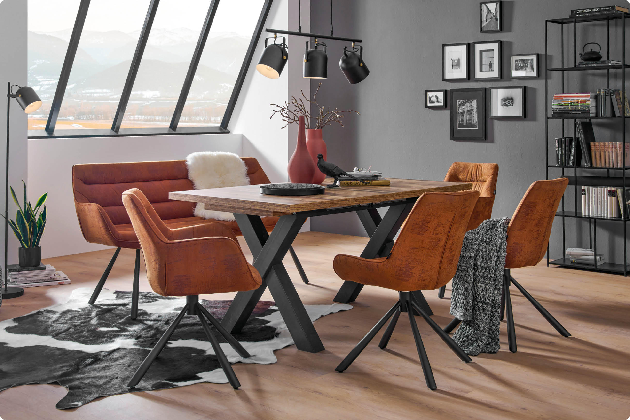 Esszimmer mit braunen Stühlen und Sofa um einen Holztisch mit schwarzen Metallbeinen auf hellem Holzboden vor großen Fenstern mit Bergblick