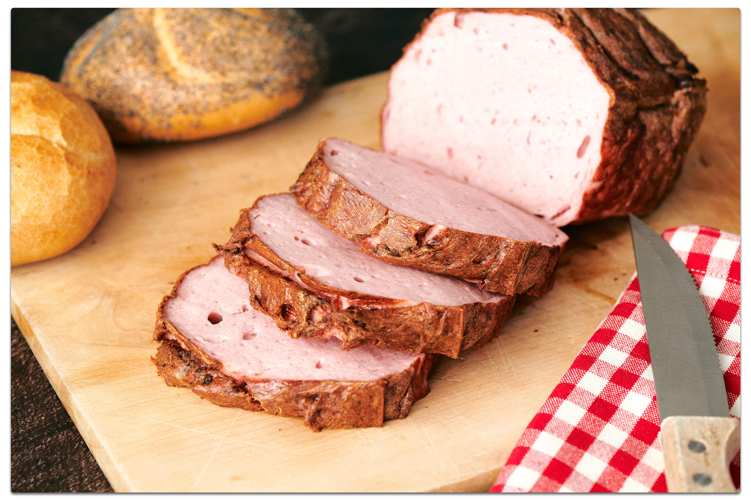 Auf einem Schneidebrett liegender, geschnittener Fleischkäse, im Hintergrund 2 Kaiserbrötchen, mit einem Messer, perfekt für die Oktoberfestfeier zu Hause.
