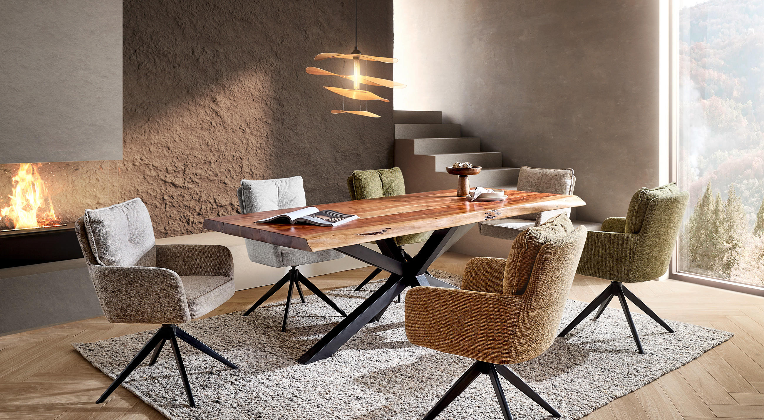 Esszimmer mit massiver Holztisch‑Platte auf schwarzem X‑Gestell, umgeben von sechs gepolsterten Drehstühlen in Beige‑ und Grün­tönen auf hellgrauem Teppich; links offener Kamin, rechts großes Fenster mit Blick ins Tal und dezente Wandleuchten