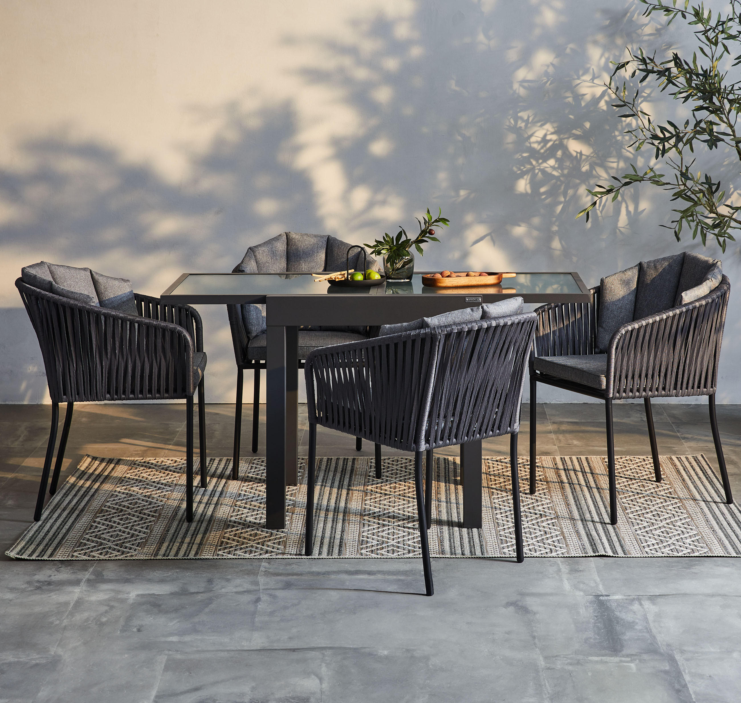 Outdoor-Dining-Set mit dunklen Stühlen, Glastisch und Deko auf Teppich, vor Wand mit Pflanzenschatten