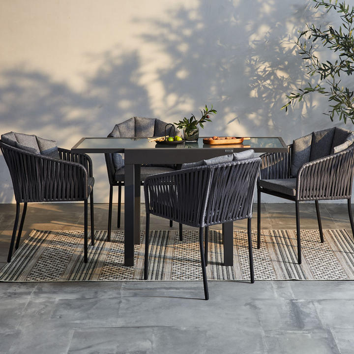 Outdoor-Dining-Set mit dunklen Stühlen, Glastisch und Deko auf Teppich, vor Wand mit Pflanzenschatten