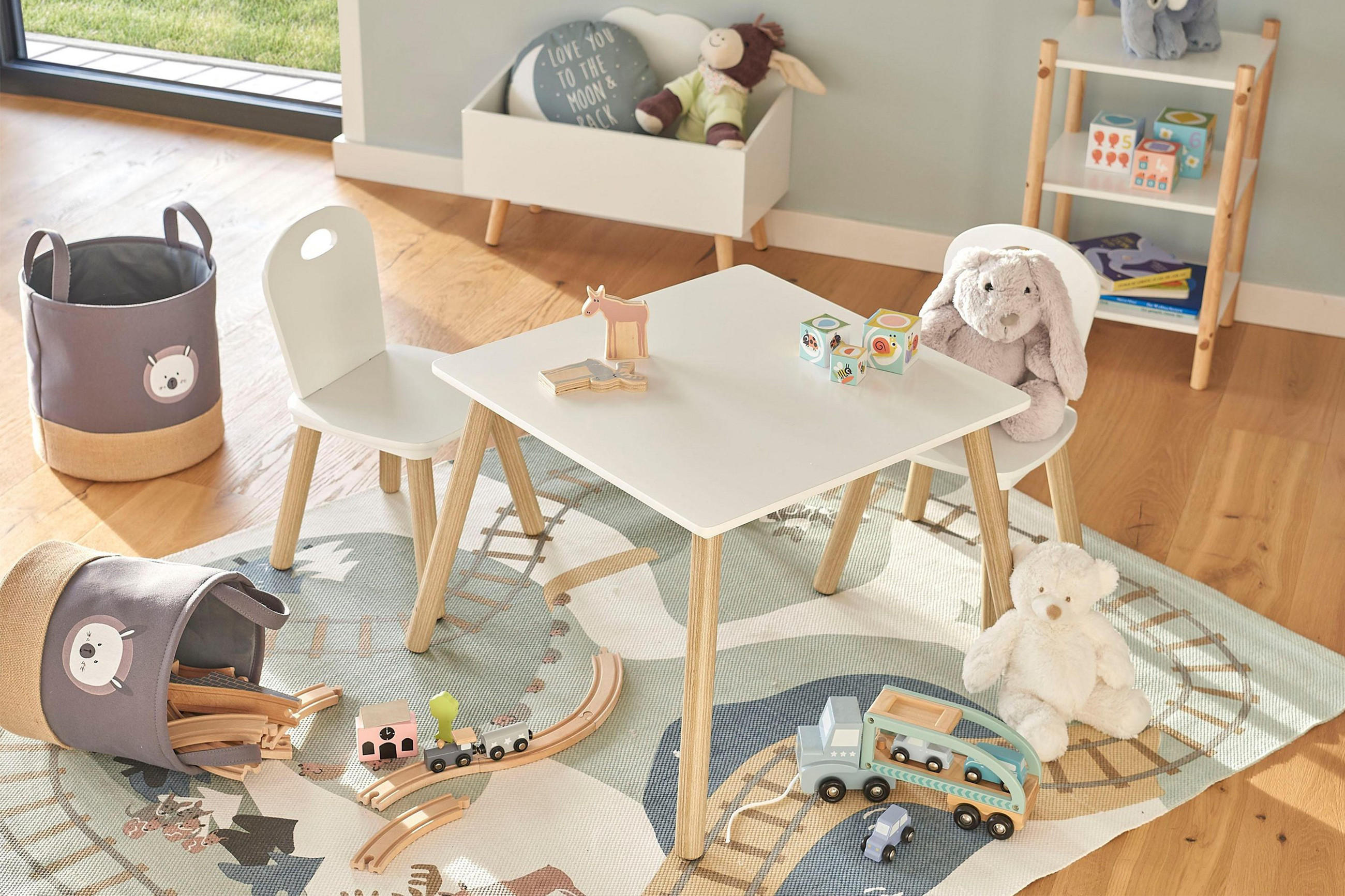 In einer Spielecke mit Kindertischgruppe, sitz ein Stoffhase, gemeinsam mit Kinderteppich und Spielzeugautos aus Holz, sowie Körben und Regalen mit weiteren Spielwaren.