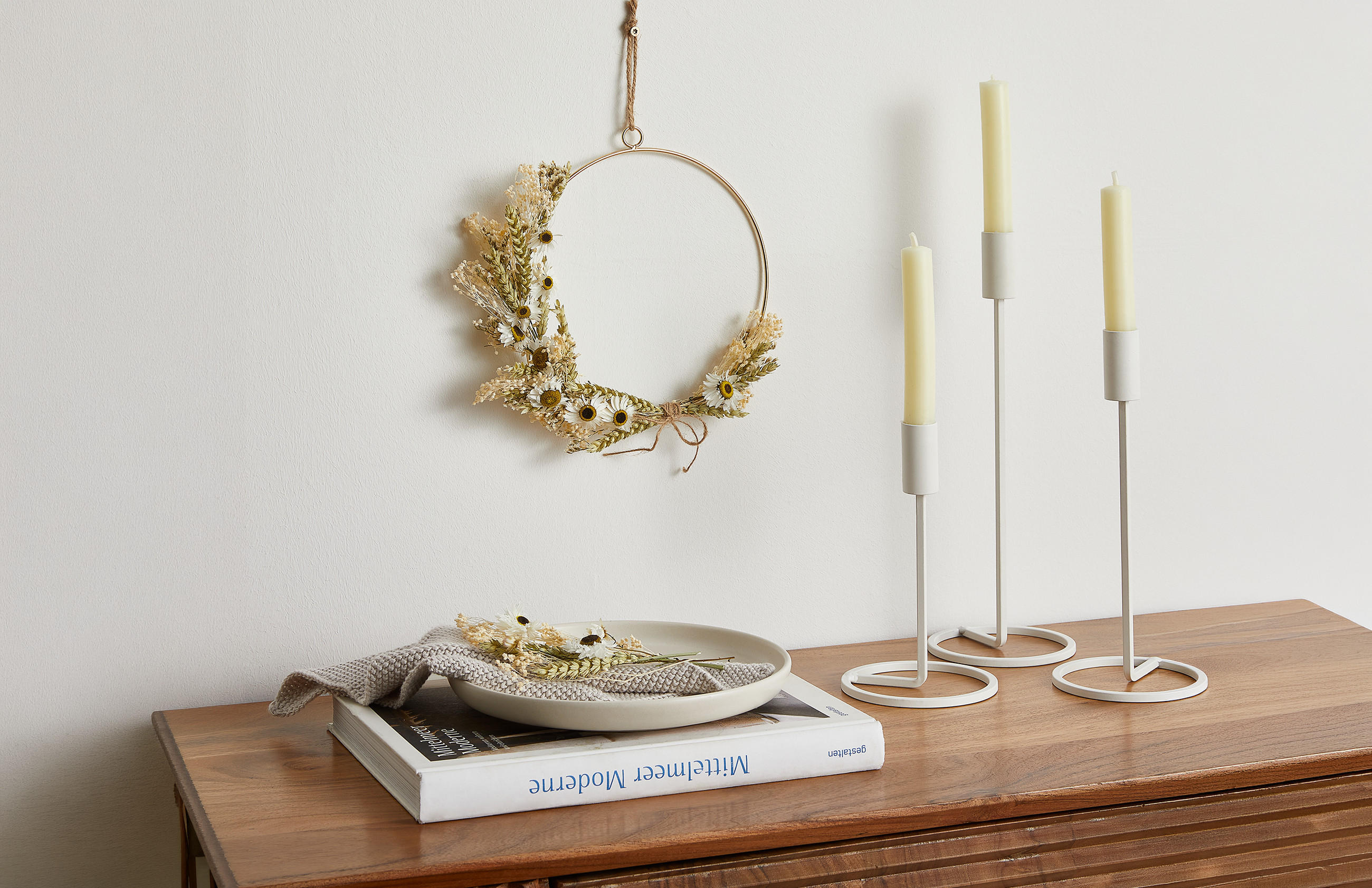 Trockenblumenkranz an der Wand über einem Holzsideboard mit Kerzenständern, einem Buch und einem Teller mit Stoffserviette.