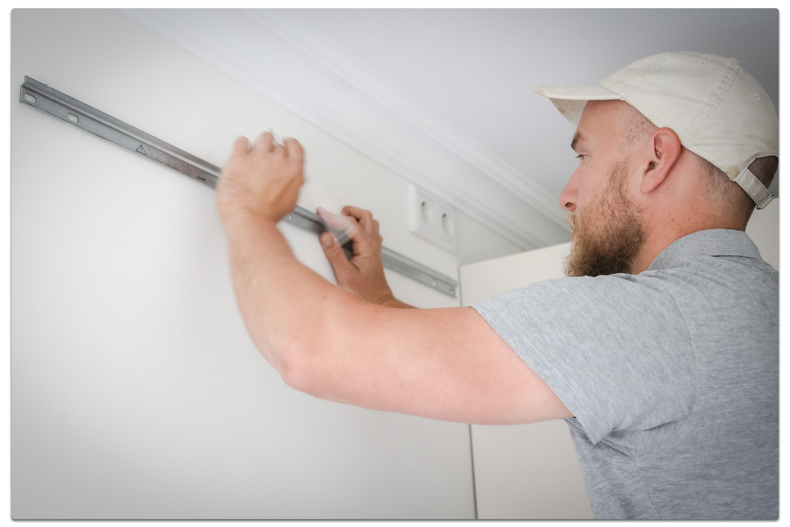 Mann mit kurzer grauer Kleidung und einer weißen Kappe, der eine Metallleiste an einer Wand anbringt. Er hält ein Messwerkzeug in der Hand.