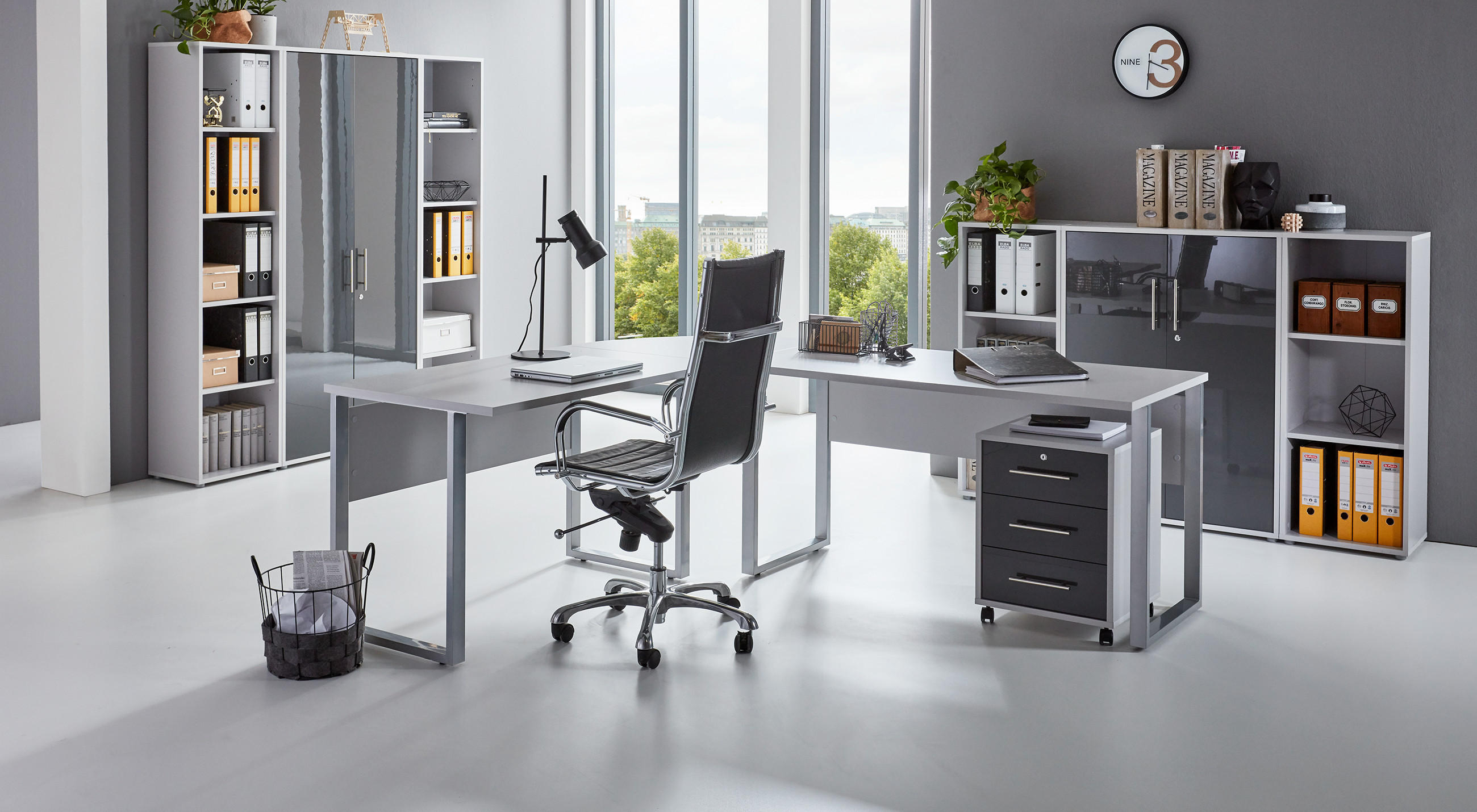 Modernes Büro mit großem Schreibtisch, Laptop, Lampe und Dokumenten, umgeben von Regalen und Aktenschränken; im Hintergrund große Fenster mit Blick ins Grüne.