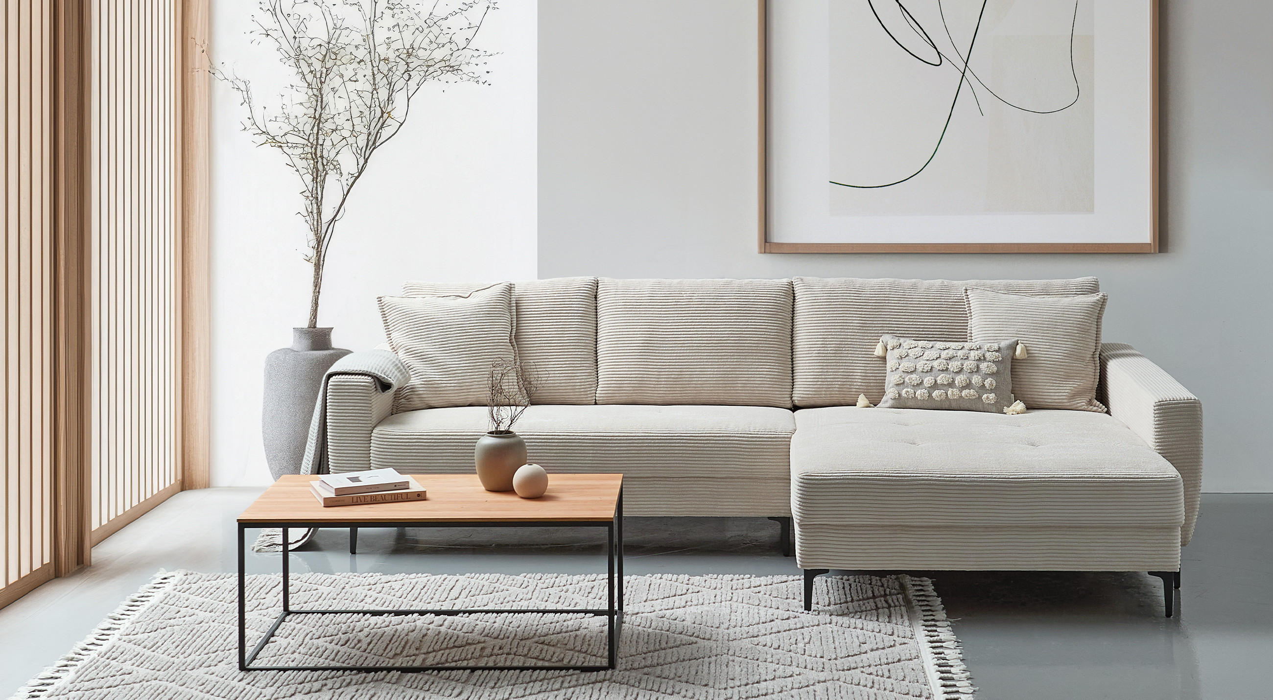 Hellbeiges Cord‑Ecksofa mit Chaiselongue rechts, flacher Holztisch mit schwarzem Metallgestell auf gemustertem Teppich; neben dem Sofa große Vase mit getrockneten Zweigen, helle Holzlatten‑Trennwand links und abstraktes Bild an der Wand.