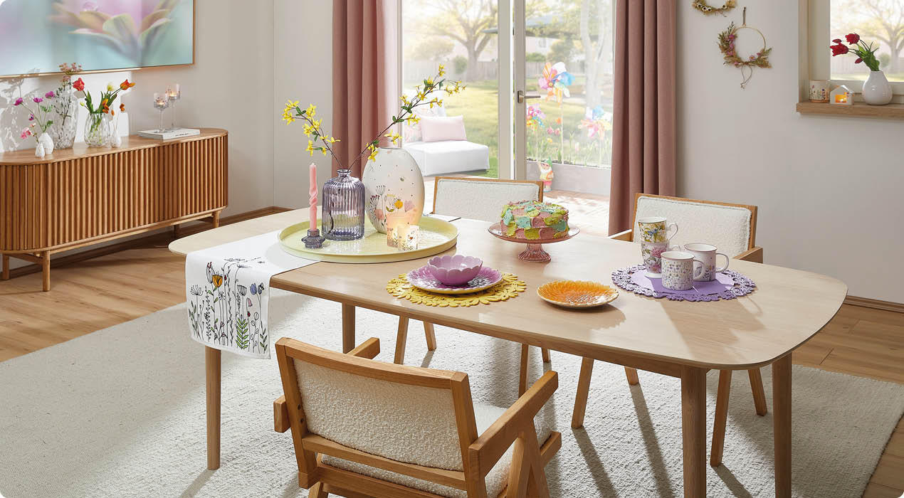 Esszimmer mit hellem Holztisch, vier gepolsterten Stühlen, dekorativen Tischsets, Vasen mit Blumen und großem Fenster mit Blick in den Garten