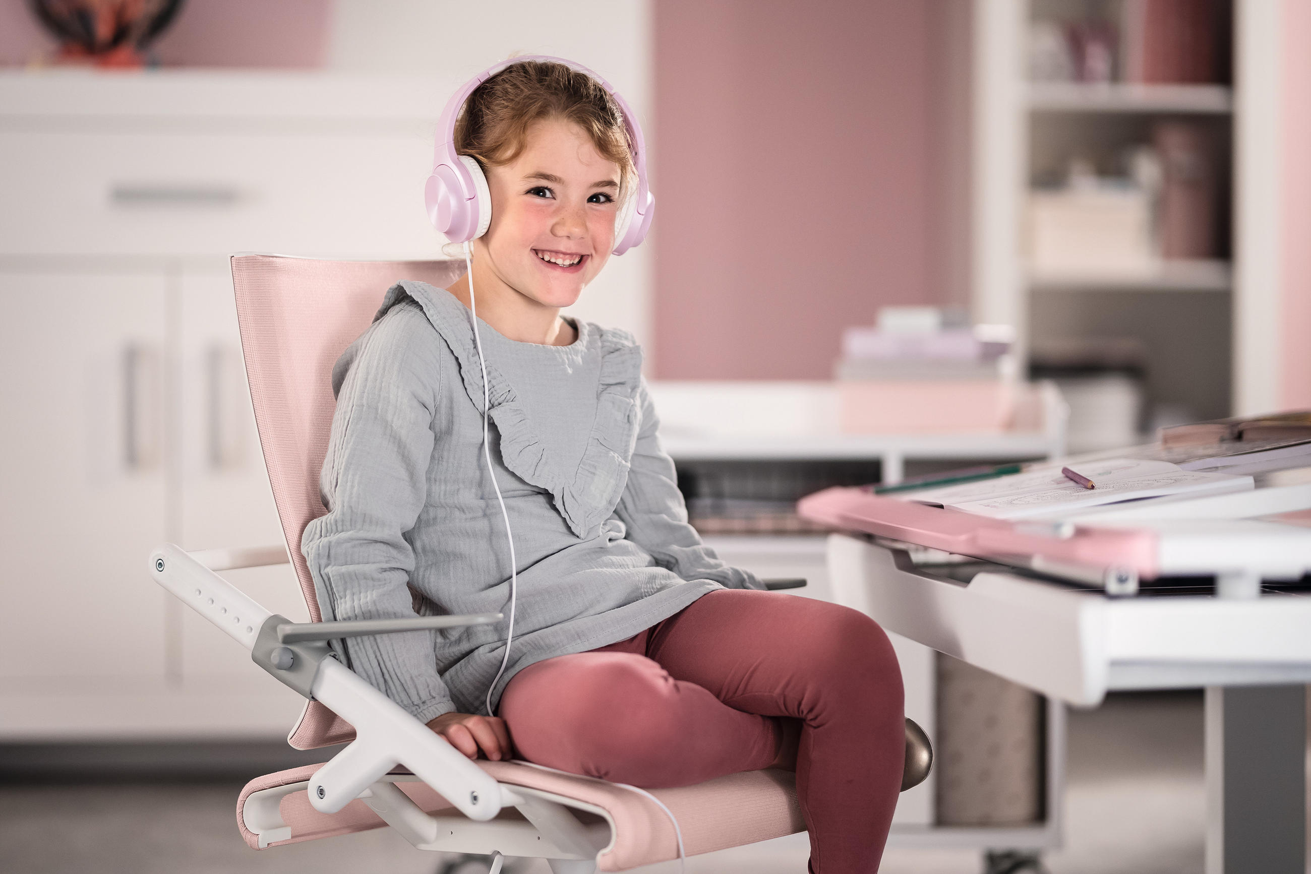 Mädchen sitzt am Schreibtisch in rosa-weißem Schreibtischstuhl mit Kopfhörern und grinst