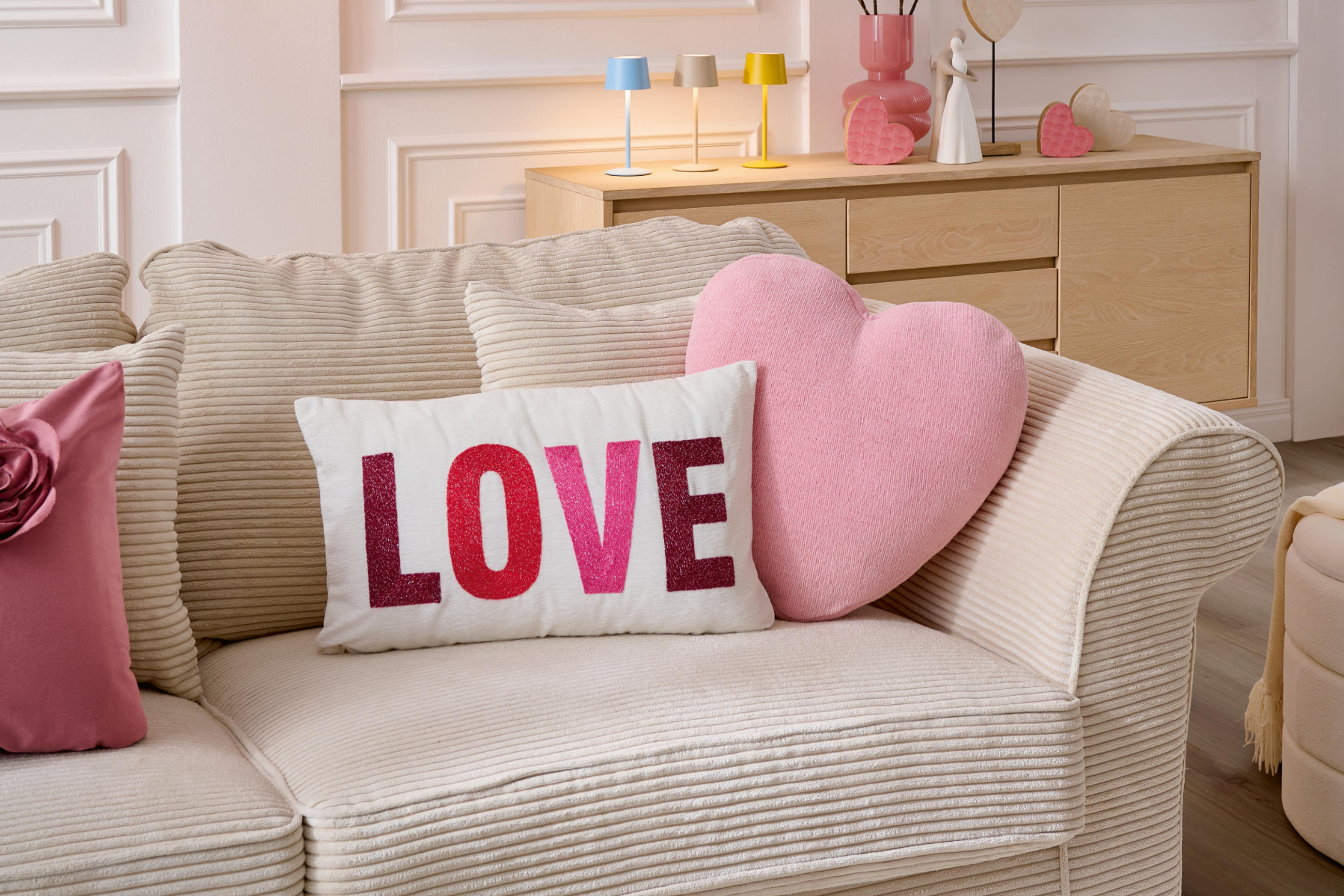 Weiße Couch mit rosa und weißen Heimtextilien, Love Kissen und Dekoherzen im Hintergrund auf einer braunen Holzkommode