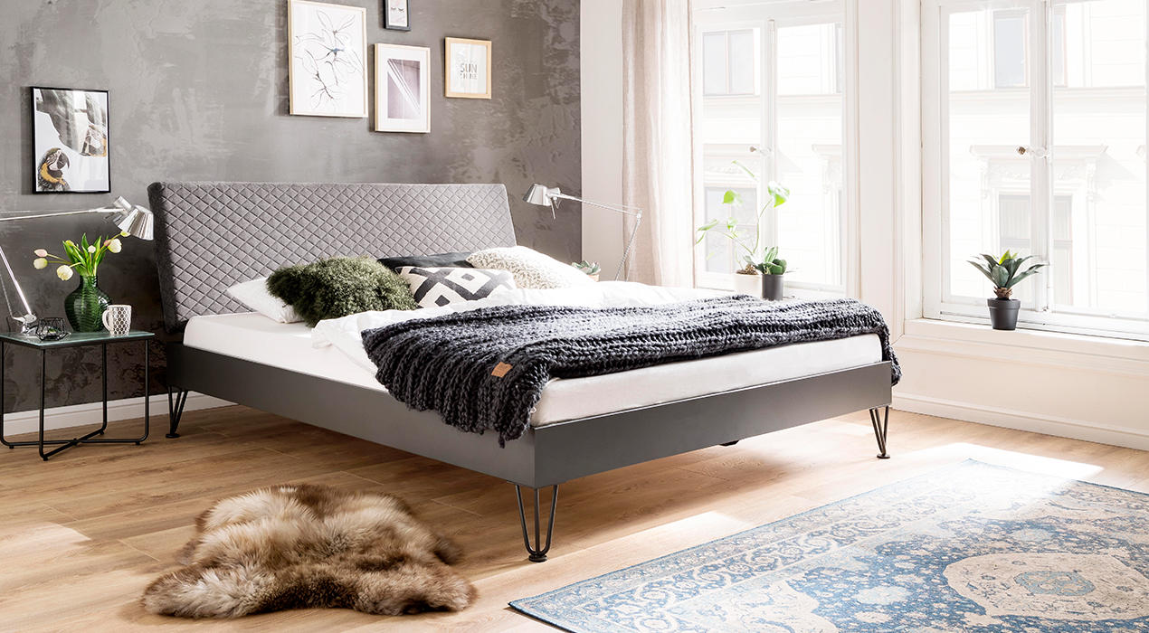 Graues Bett auf Metallfüßen mit Steppung im Kopftei.