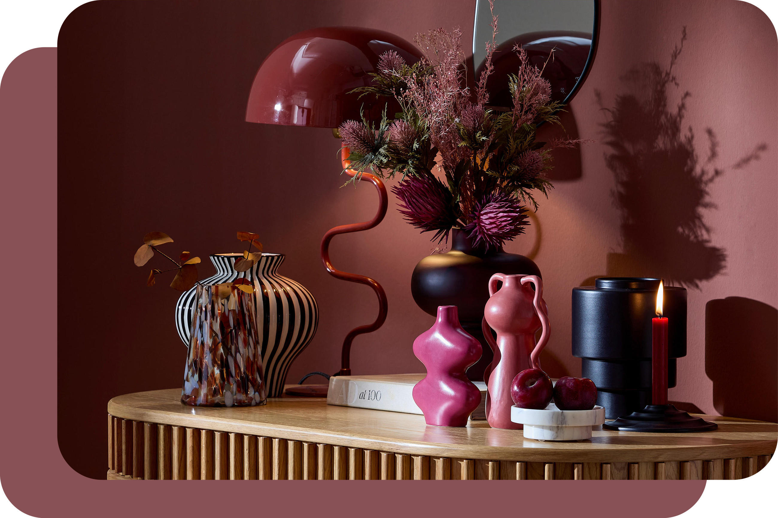 Dekoration auf Holztisch vor burgunderroter Wand: rote Lampe, Trockenblumen, rosa Vasen, rote Äpfel und kleiner Spiegel.