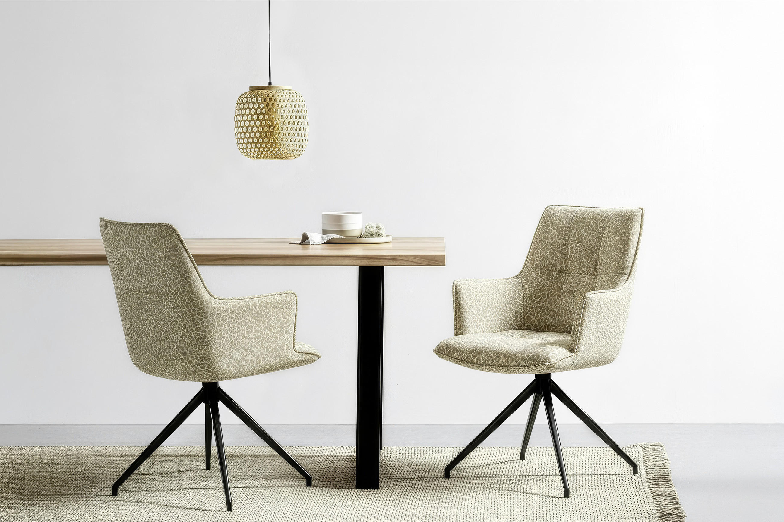 Minimalistisches Esszimmer-Setting mit BELLUTI Holztisch auf schwarzem Metallgestell und zwei gepolsterten Drehstühlen in hellem Musterstoff; darüber eine pendelnde Designleuchte vor weißer Wand.