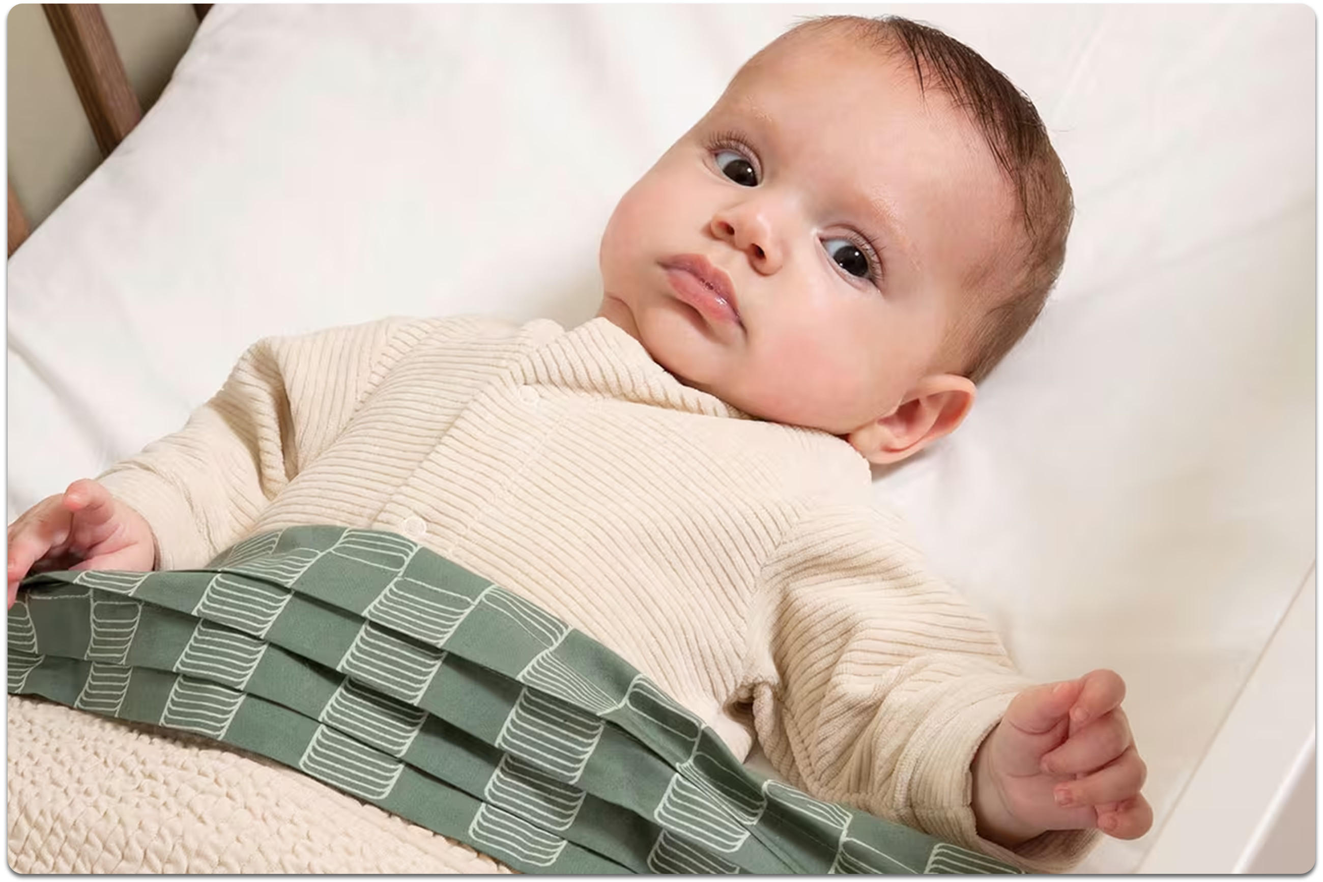 Baby mit braunen Haaren liegt in einem Babybett mit beigem Body und grün-karierter Decke und blickt in die Kamera.