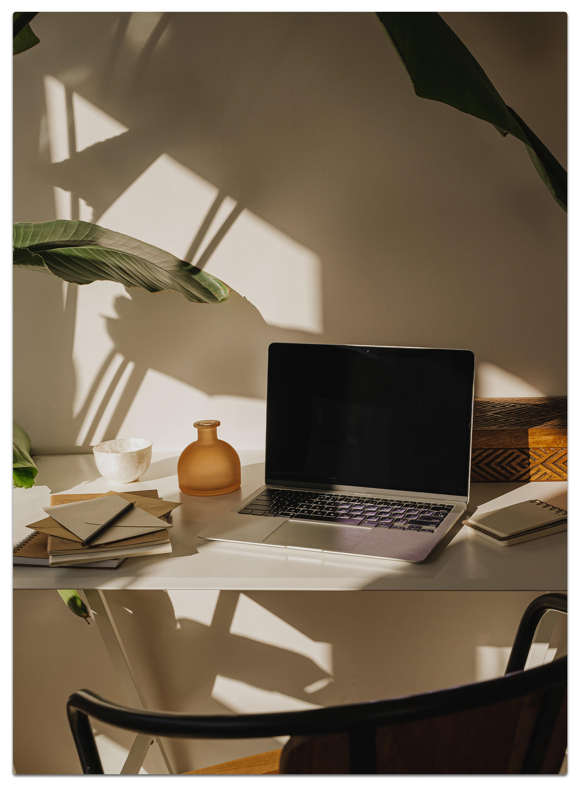 Heller Arbeitsplatz mit Laptop, Notizbüchern und grüner Blattschatten an der Wand.