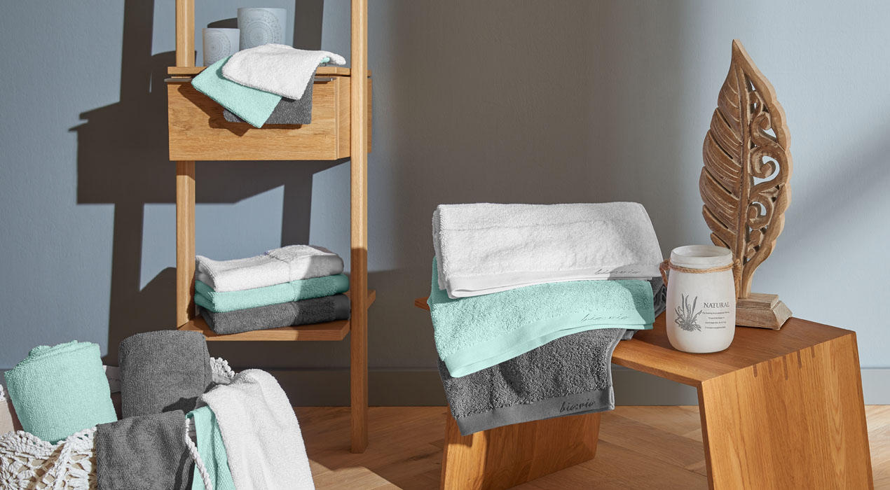 Farbenfrohe Heimtextilien in Türkis und Grau, darunter Handtücher und Decken, dekorativ arrangiert auf Holzregalen