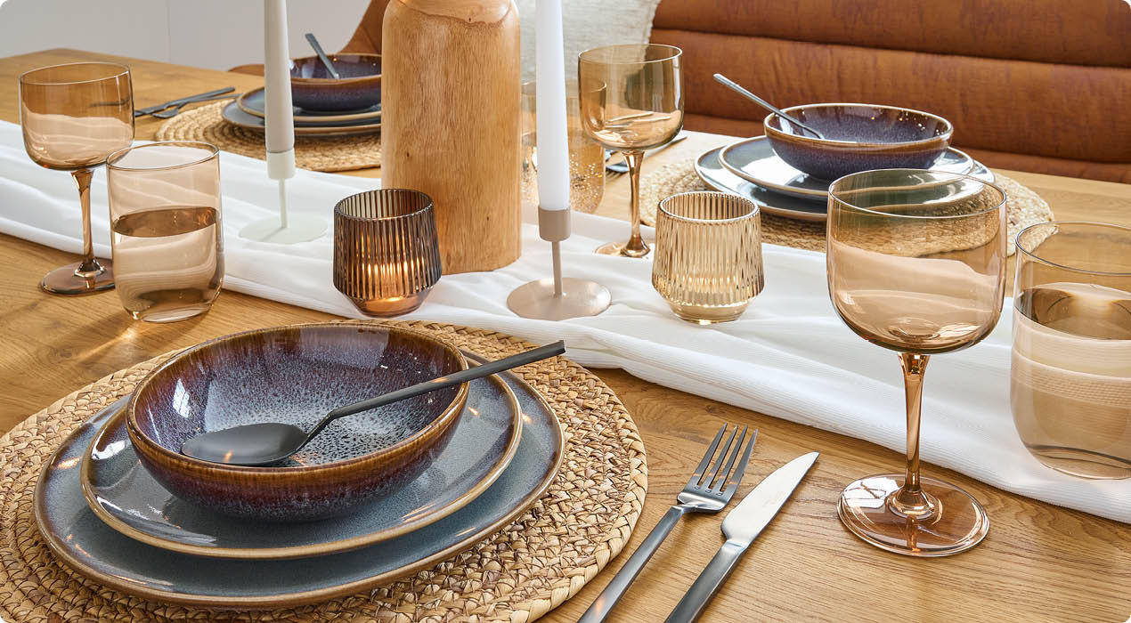 Esstisch mit geflochtenem Platzset, blau-grauen Keramiktellern, Besteck, bernsteinfarbenen Gläsern, Kerzenhaltern und einer Holzvase als Dekoration