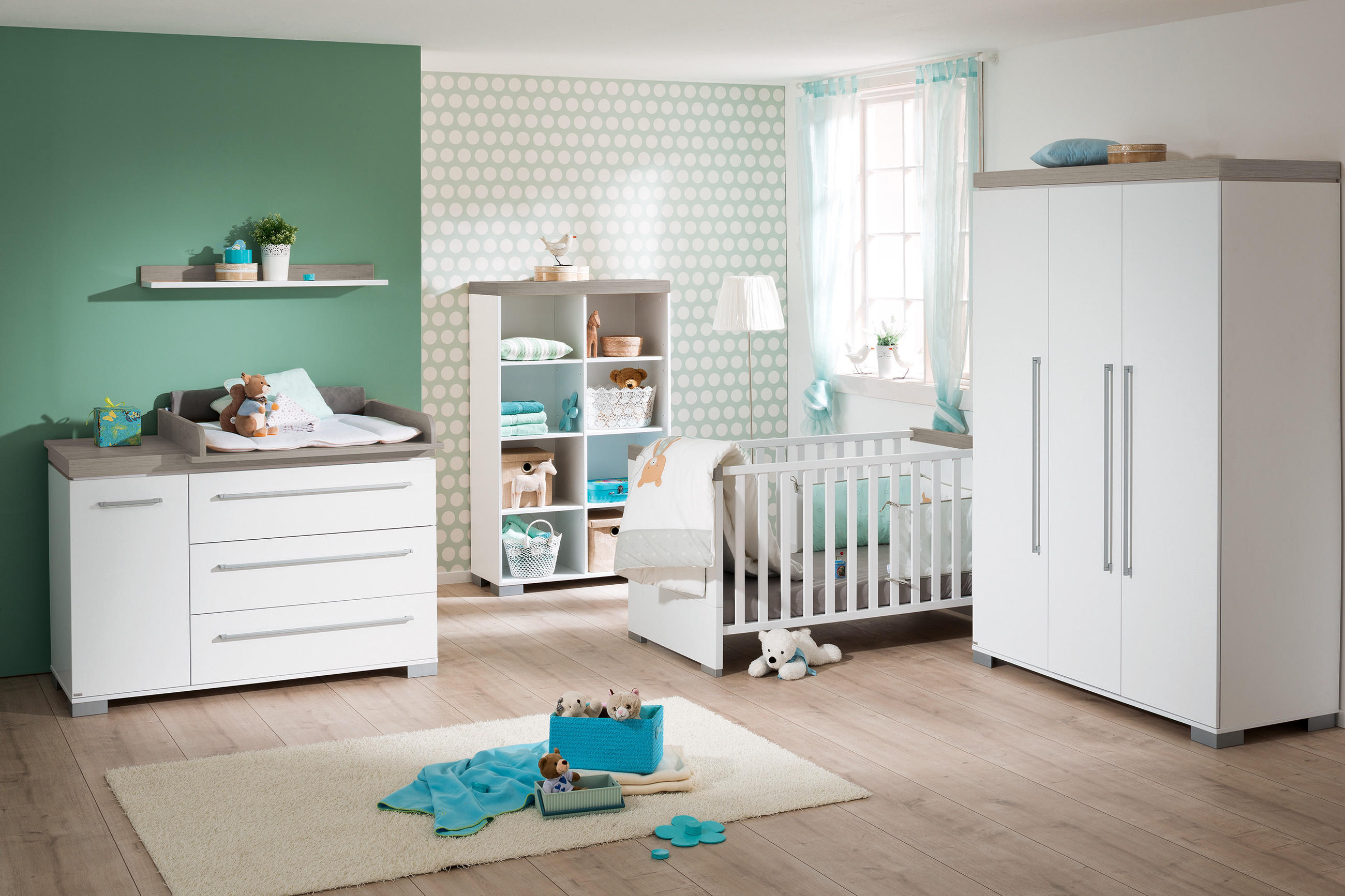 Kinderzimmer mit Wickelkommode, Regal, Kinderbett und Kleiderschrank in hellem Holz und Weiß vor grüner Wand.