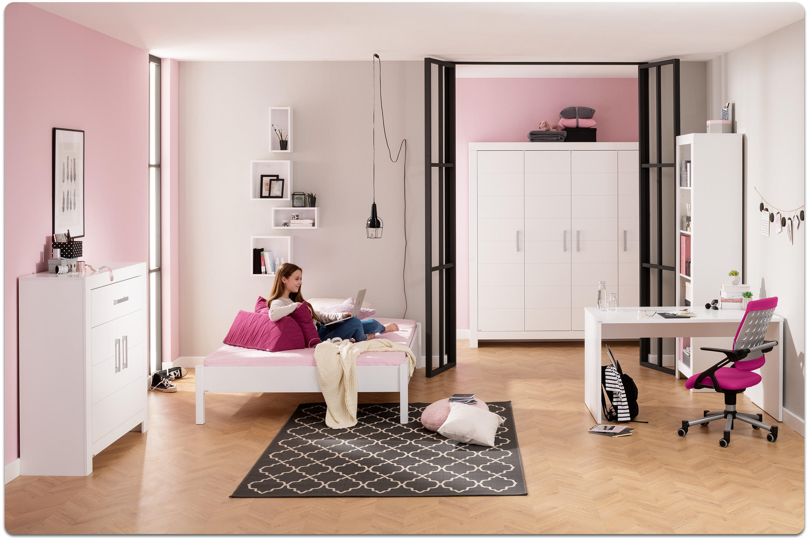 Jugendliches Mädchen sitzt auf einem weißen Bett mit pinken Kissen in einem hellen, modernen Jugendzimmer mit Schreibtisch, Stuhl, Schrank und Teppich.