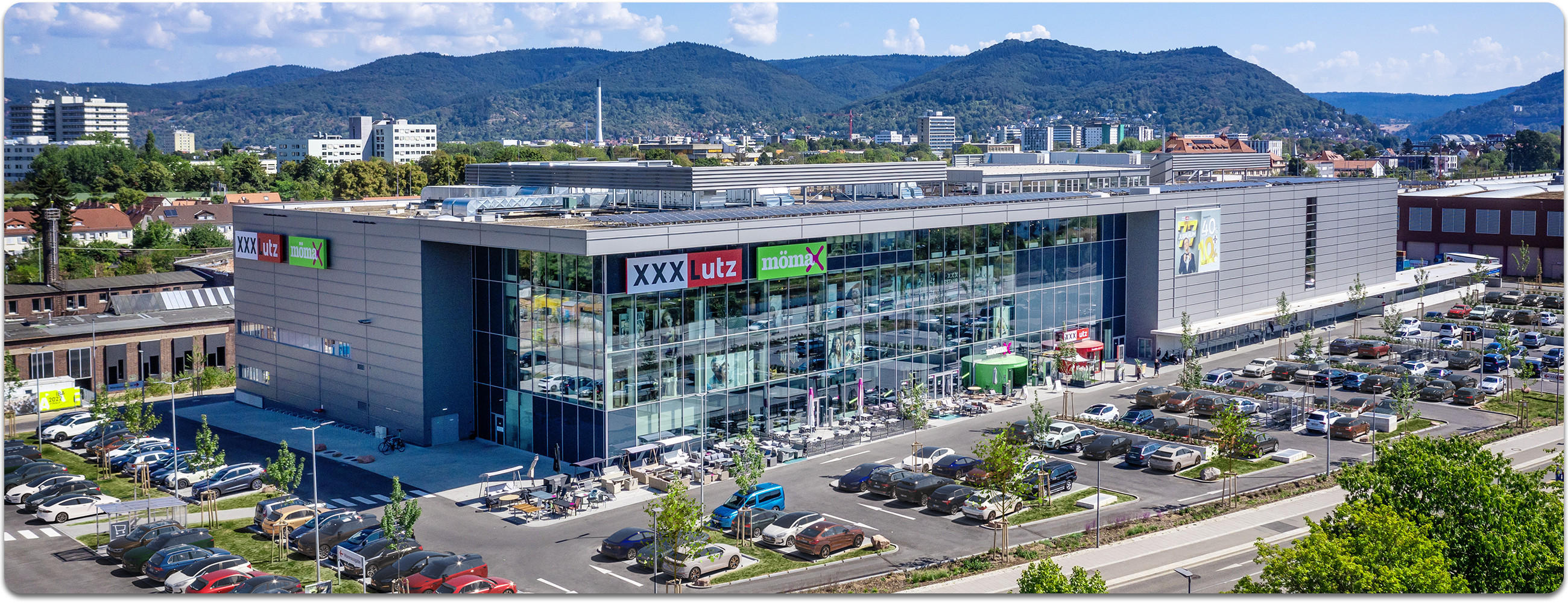 Luftaufnahme der XXXLutz-Filiale in Heidelberg mit großem Parkplatz voller Autos, breiter Straße im Vordergrund und Stadtbebauung unter blauem Himmel im Hintergrund.