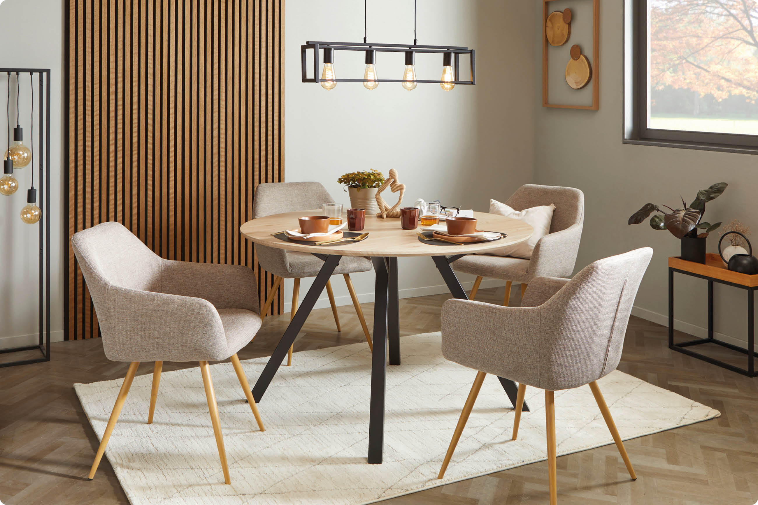 Esszimmer mit rundem Holztisch, vier gepolsterten Stühlen mit Holzbeinen, Teppich, Pflanzen und Wanddeko