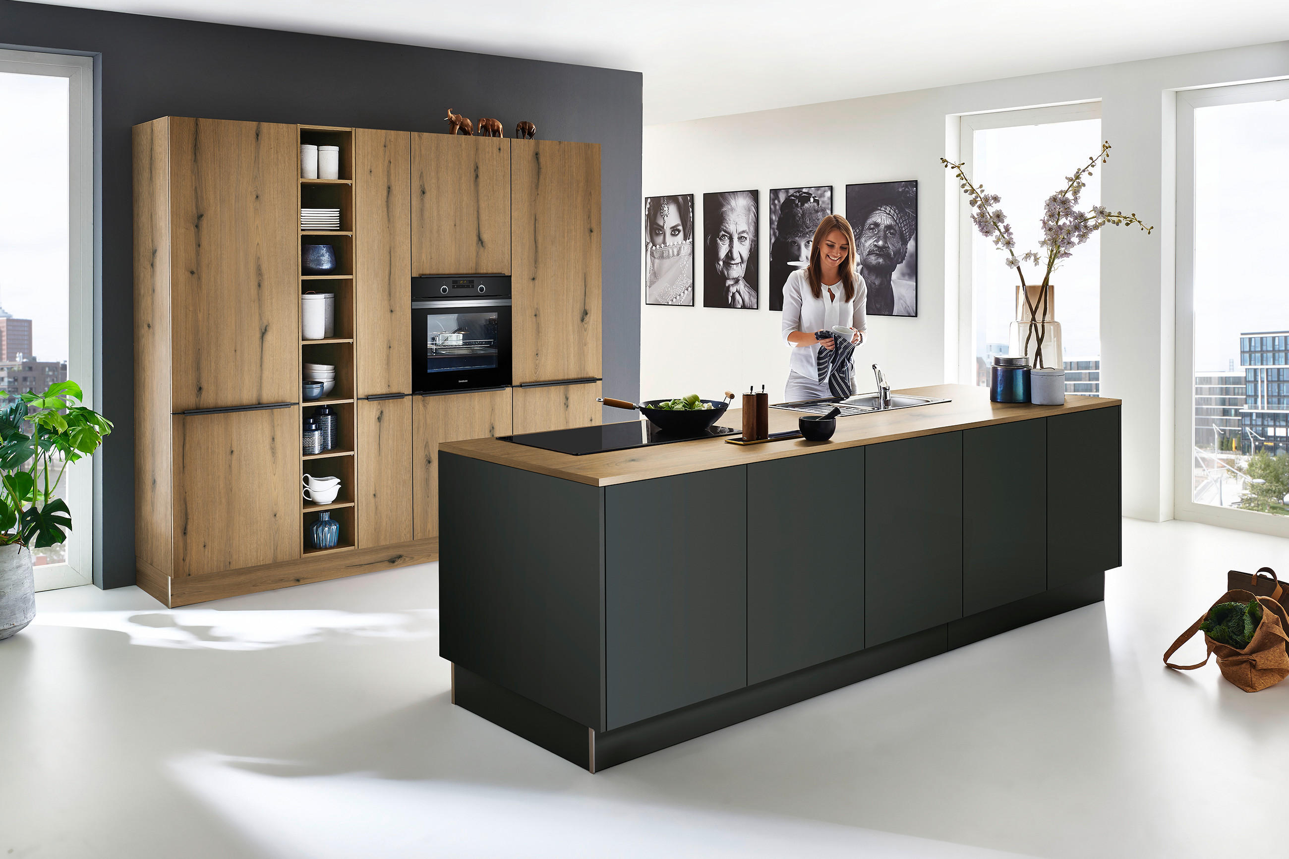 Helle, moderne Küche in Naturtönen: Küchenschrank in warmem Eiche-Holzdekor mit offenen Regalfächern und integriertem Backofen, dazu eine Kücheninsel mit dunklen, matten Fronten und Holzarbeitsplatte.