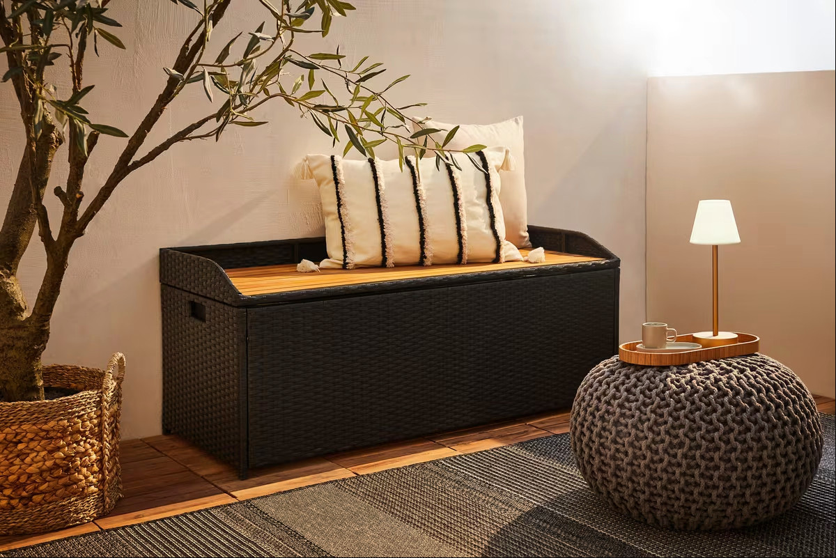 schwarze Kissenbox auf Holzboden und ein geflechteter Aufbewahrungskorb vor einem grauen Outdoorteppich mit einem grauen Sitzkissen