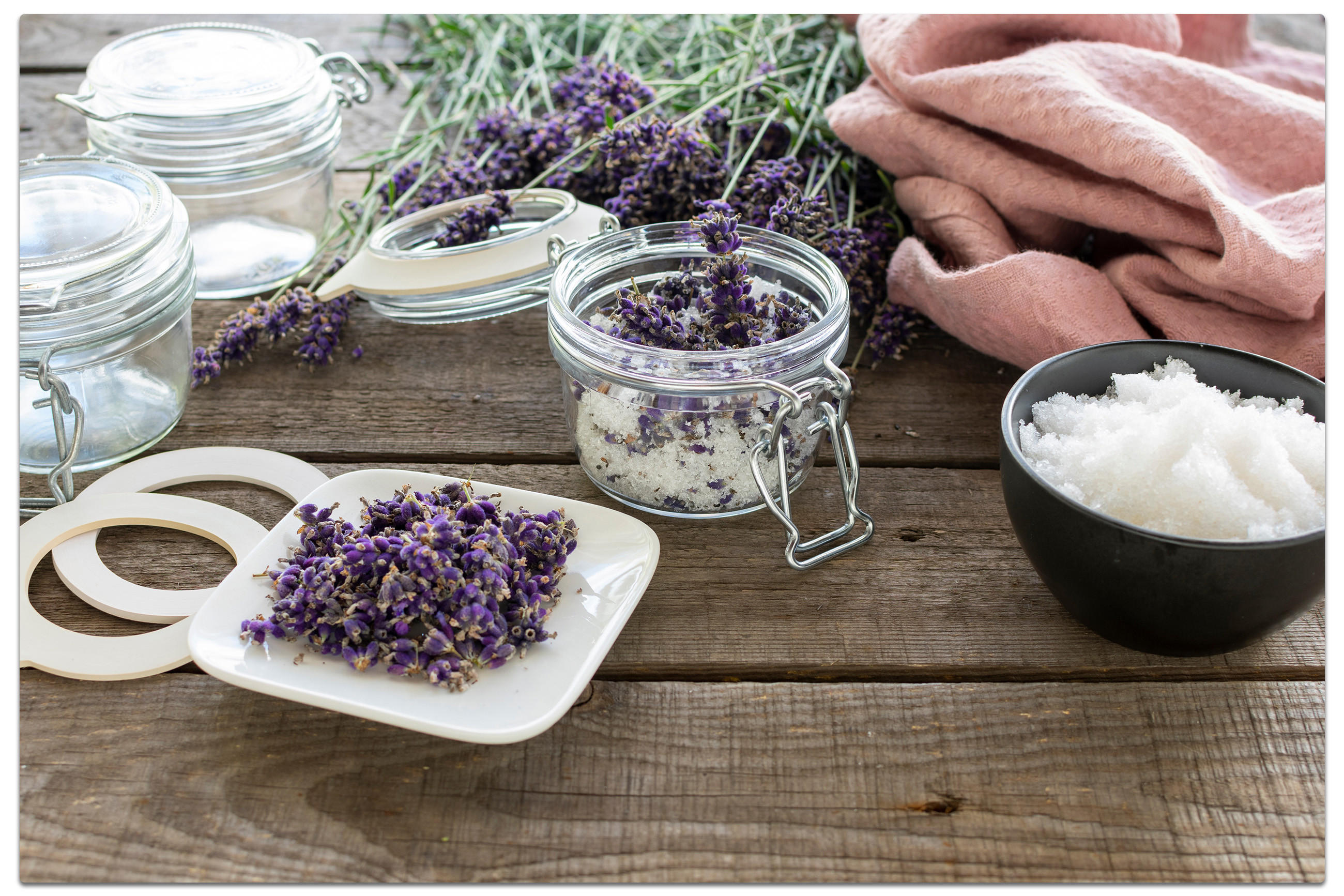 Rustikaler Holztisch mit Gläsern, getrocknetem Lavendel und Salz. Im Hintergrund frischer Lavendel und rosa Stoff