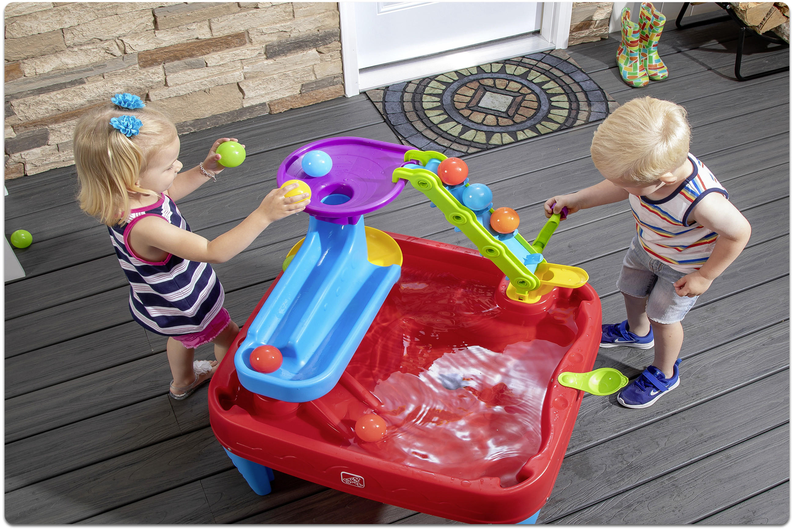 Zwei Kleinkinder spielen an einem roten Wasserspieltisch mit blauer Rutsche und grünem Kran auf einer Holzterrasse.
