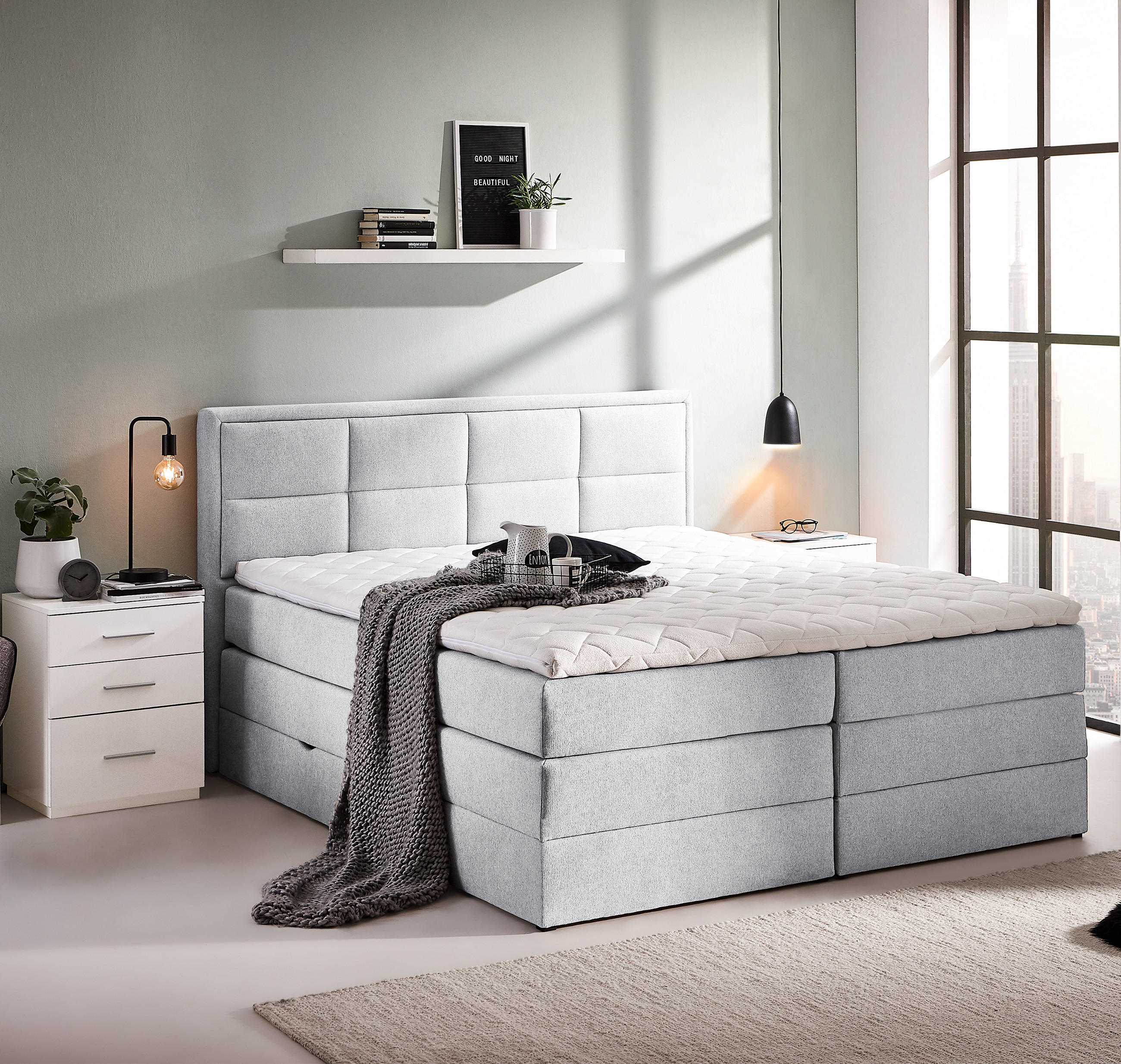 Modernes Schlafzimmer mit grauem Boxspringbett, Nachttisch, Stuhl, Pflanzen und großen Fenstern.
