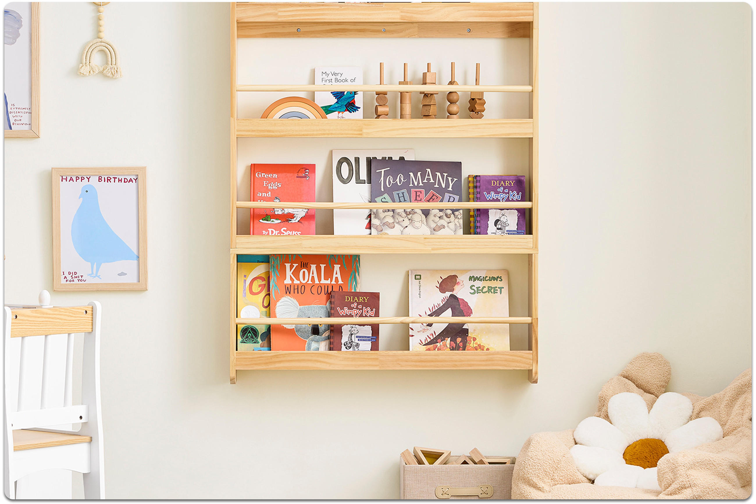 Holzregal mit Kinderbüchern und Holzspielzeug an weißer Wand über einem Sitzbereich mit Kissen in Form einer Blume und einem Korb.