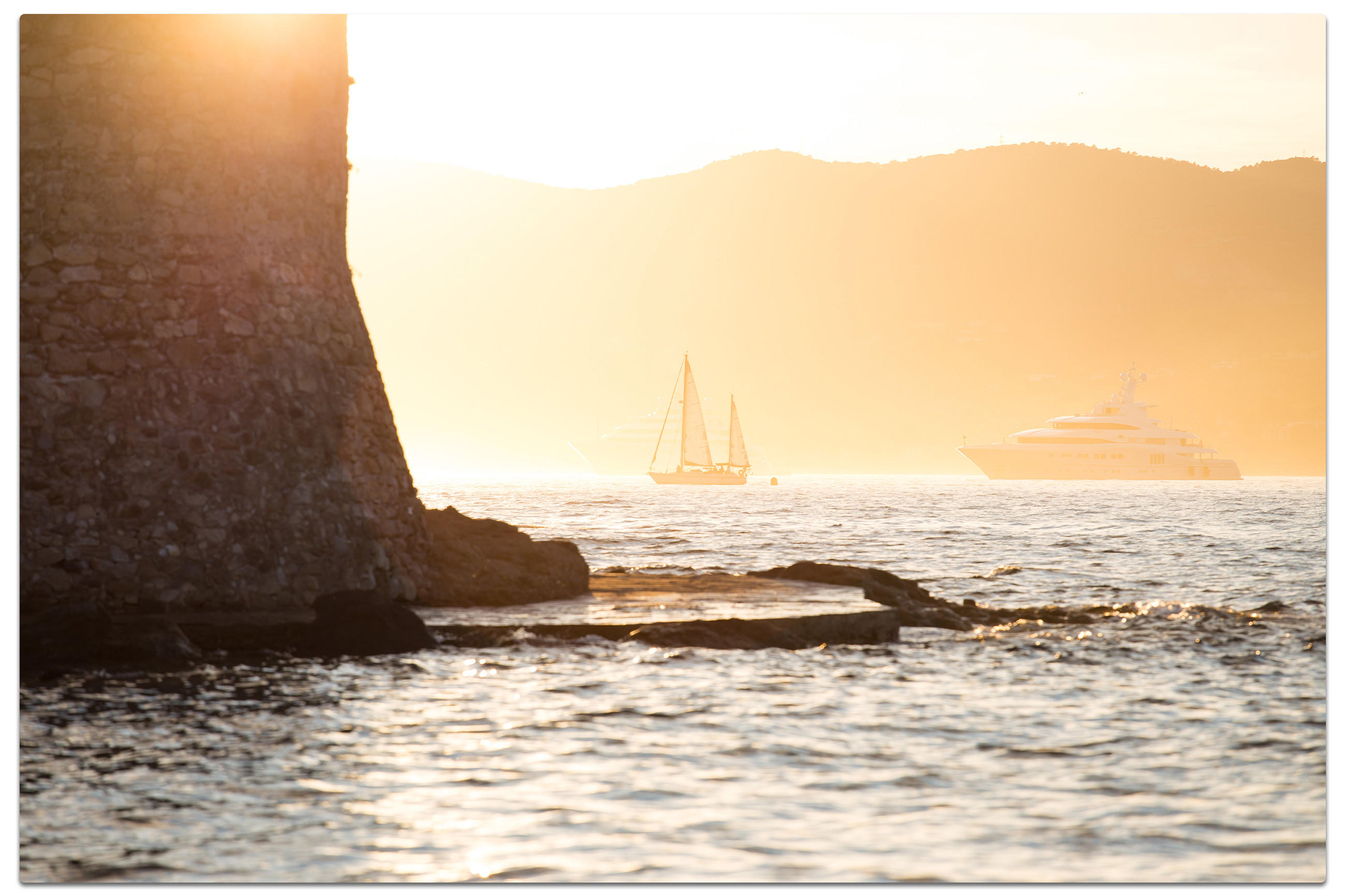 Sonnenuntergang über dem Meer mit Segelboot, Yacht und Steinstruktur im Vordergrund.