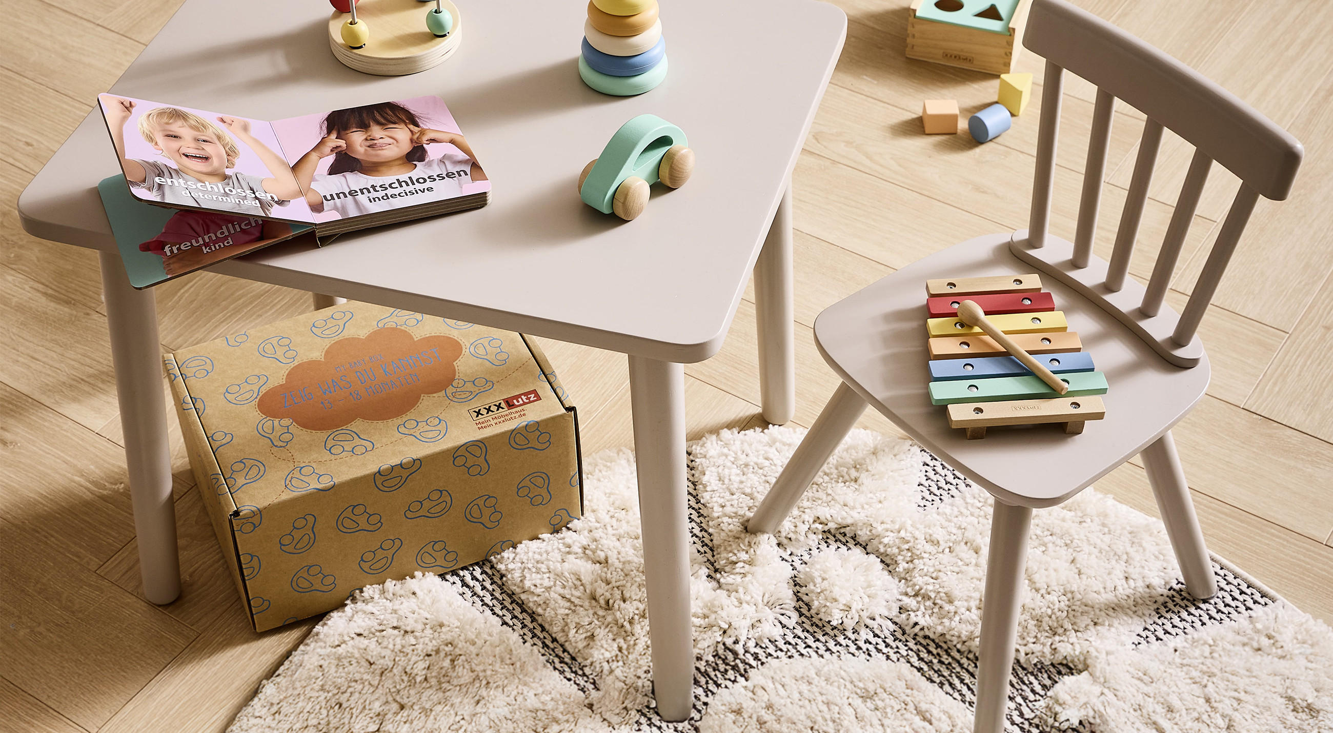 Kinderzimmer-Set mit kleinem Tisch und Stuhl, Holzspielzeug und Xylophon; auf dem Tisch ein Kinderbuch, daneben eine XXXLutz My Baby Box auf hellem Holzboden.