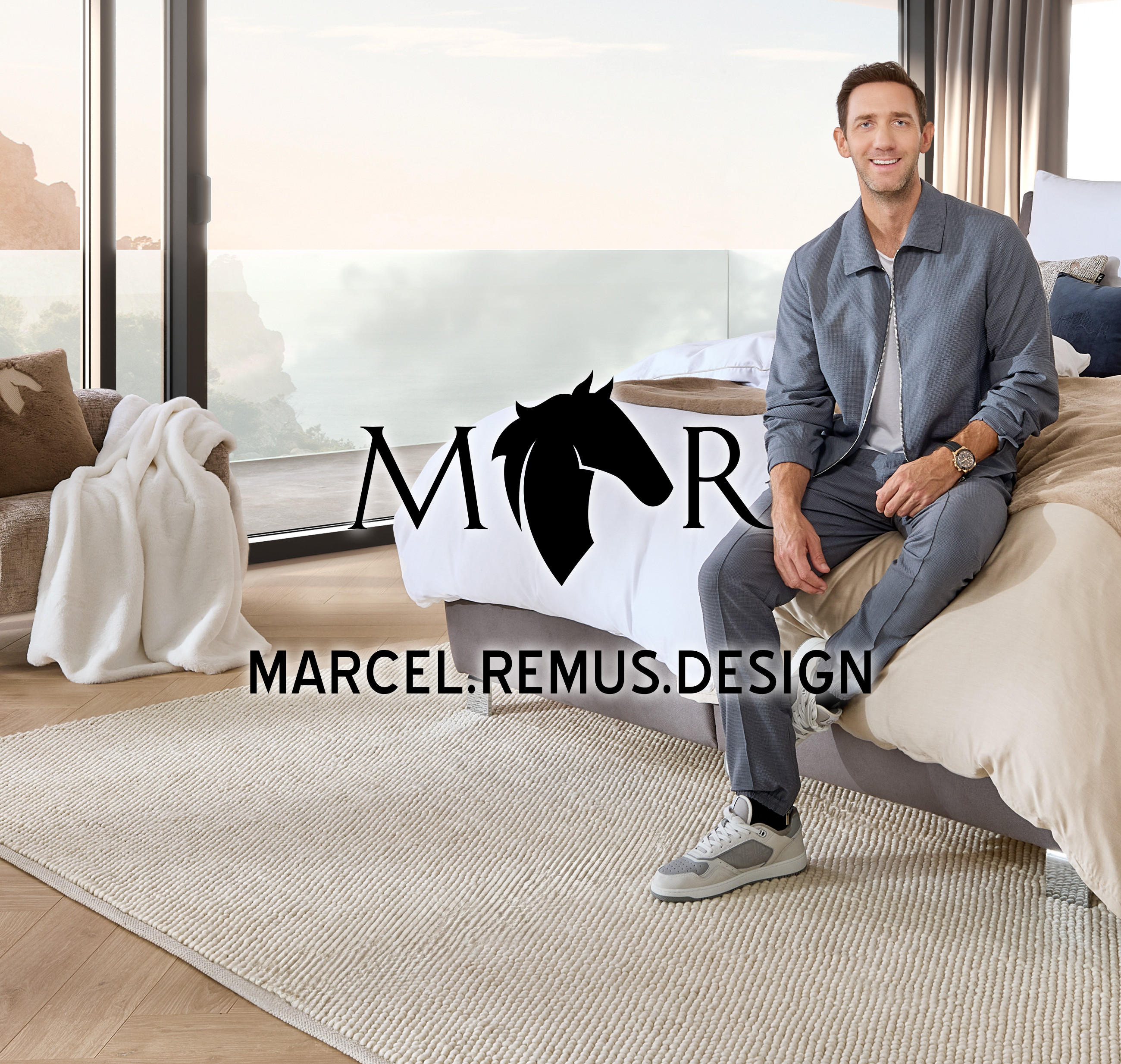 Modernes Schlafzimmer mit großem Bett in Beige- und Brauntönen, dekoriert mit Kissen und Tagesdecke. Person sitzt am Bettrand, links Sessel mit Decke, im Hintergrund Fenster mit Berg- und Meerblick, mittig Logo ‚Marcel Remus Design‘