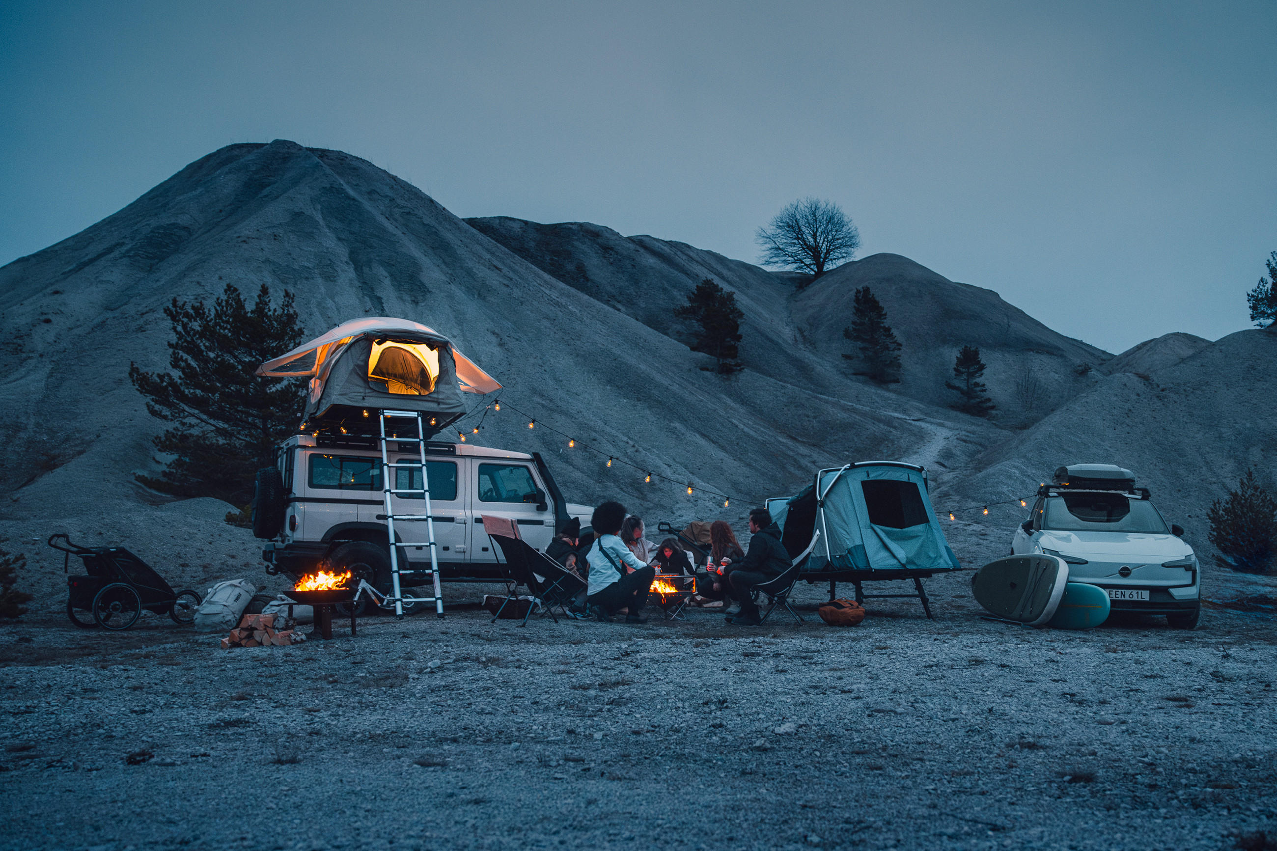 Campingplatz bei Dämmerung mit Dachzelt auf Auto, Lagerfeuer und mehreren Zelten und Ausstattung von THULE vor bergiger Landschaft
