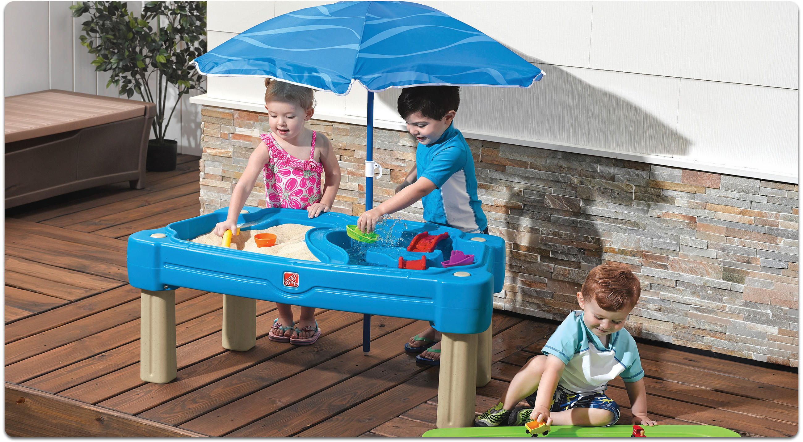 Drei Kinder spielen auf einem Balkon an einem blauen Sand- und Wasserspieltisch mit blauem Sonnenschirm vor einer weißen Wand mit Steinverkleidung