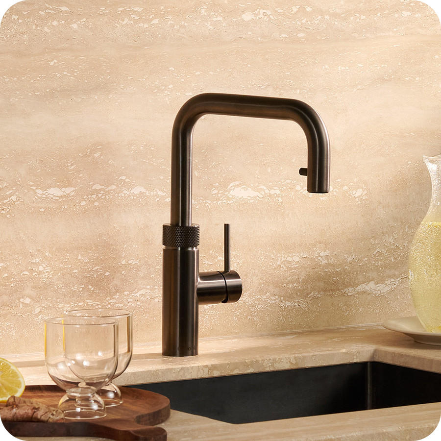 QUOOKER Küchenarmatur „Flex Square“ in dunkler Metall-Optik mit eckigem Auslauf über einer Spüle; daneben stehen zwei Gläser auf einem Holzbrett und eine Karaffe mit Getränk auf der Arbeitsplatte vor einer hellen Steinwand.