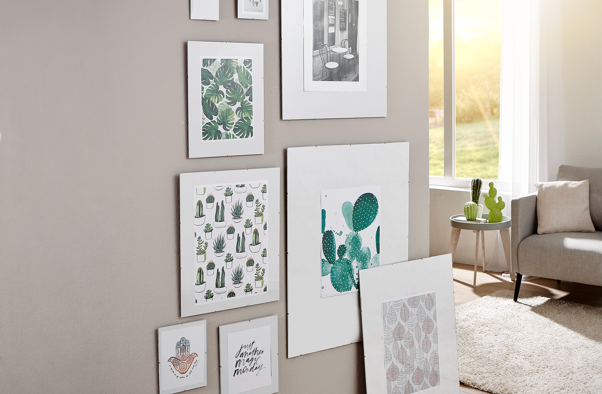 Obývacia izba, nástenné obrazy a dekorácie, stena a rámy obrazov, botanické motívy – listy, kaktusy, sukulenty, okrúhly stolík, sukulenty a dekoratívne kaktusy na odkladacom stolíku, pohovka, vankúše, svetlý koberec