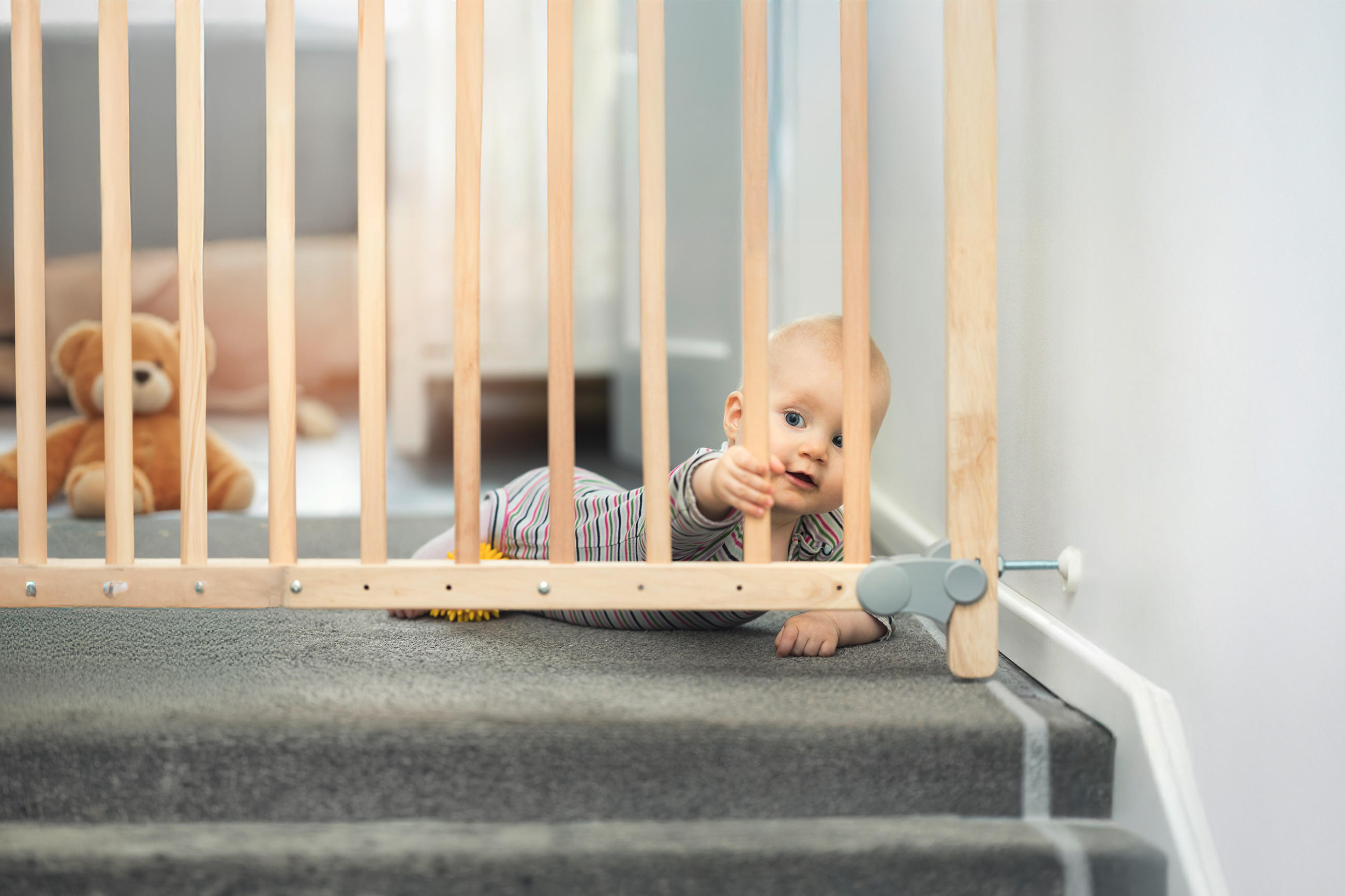 Ein Baby liegt auf dem Teppich vor einem Treppenschutzgitter und greift mit der Hand durch die Holzstäbe. Im Hintergrund sitzt ein Stofftier auf dem Boden.