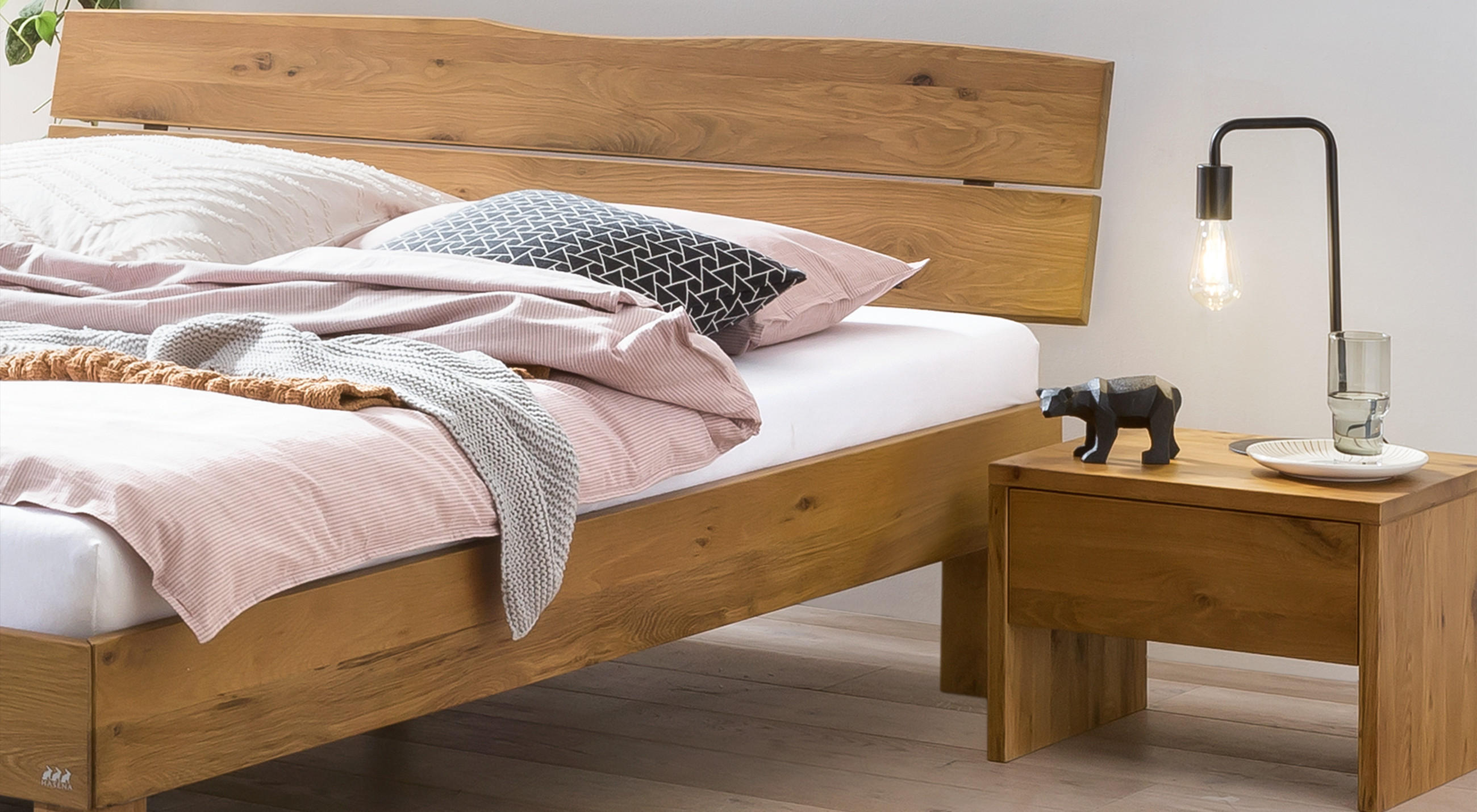 Holz‑Bett mit robustem, naturbelassenem Kopfteil und passendem Nachttisch; rosafarbene Bettwäsche, gestrickte Decke, dekoratives Kissen und Nachttischleuchte mit freiliegender Glühbirne