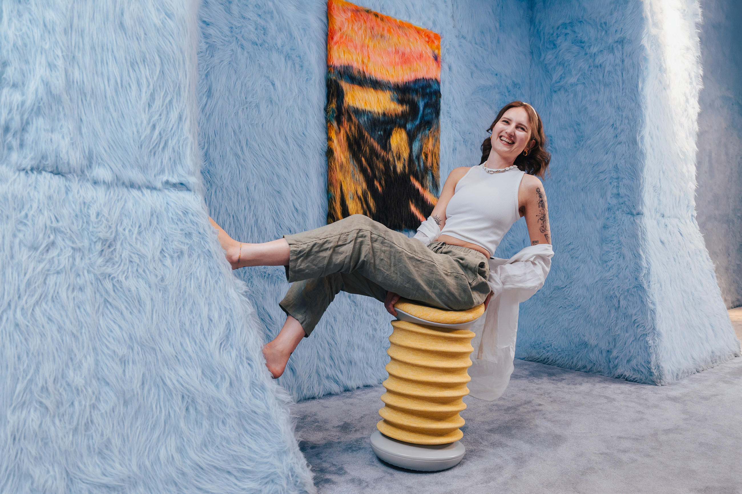 Eine Frau sitzt auf einem ergonomischen Hocker mit SITNESS Technologie von TOPSTAR, und präsentiert diesen vor einer blauen Wand mit buntem Gemälde.