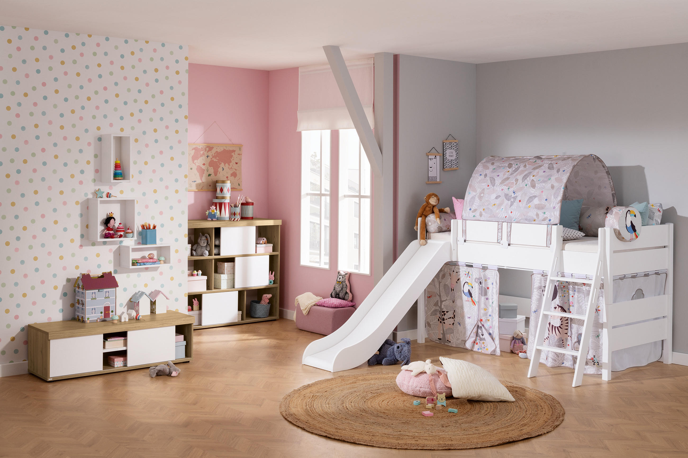 Kinderzimmer mit Hochbett und Rutsche, Teppich, Spielzeug und Regalen vor rosa und weißer Wand mit bunten Punkten.