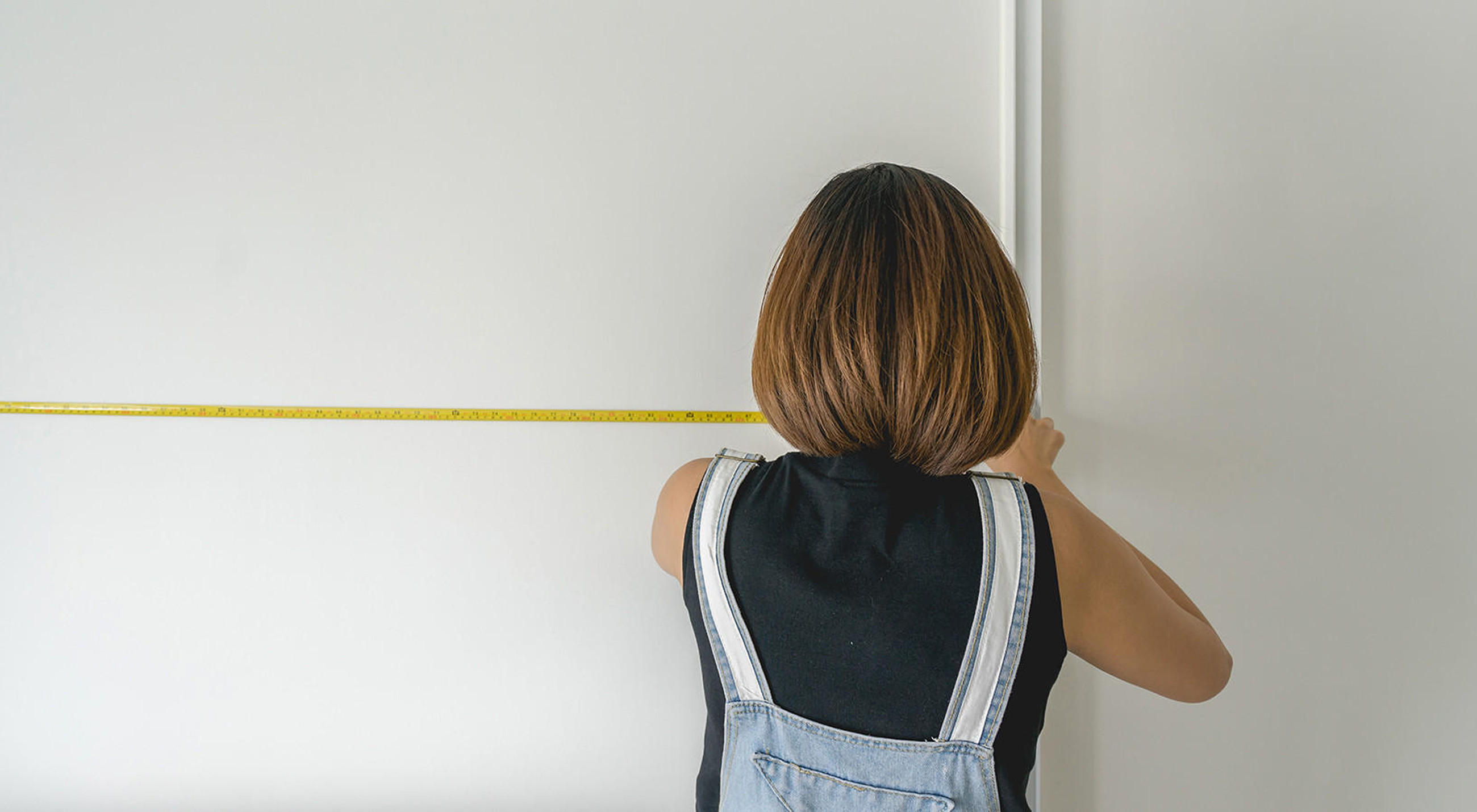 Frau mit Latzhose ist mitten in der Küchenplanung und misst eine Wand mit einem Maßband ab.