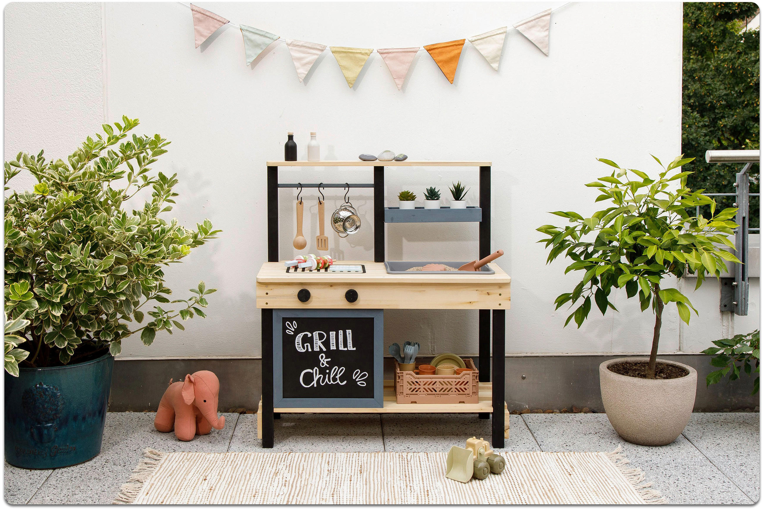 Kinderspielküche mit Grill und Kochfeld vor weißer Wand, umgeben von zwei großen Pflanzen und Spielzeug auf dem Boden.