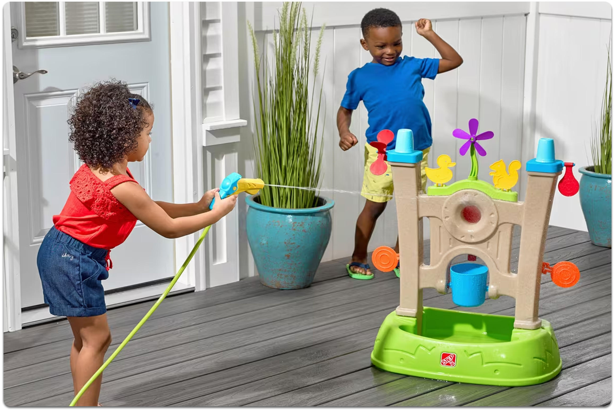 Zwei Kinder spielen auf einem Balkon mit einem Wasserspielzeug, eines hält einen Wasserschlauch, das andere springt.