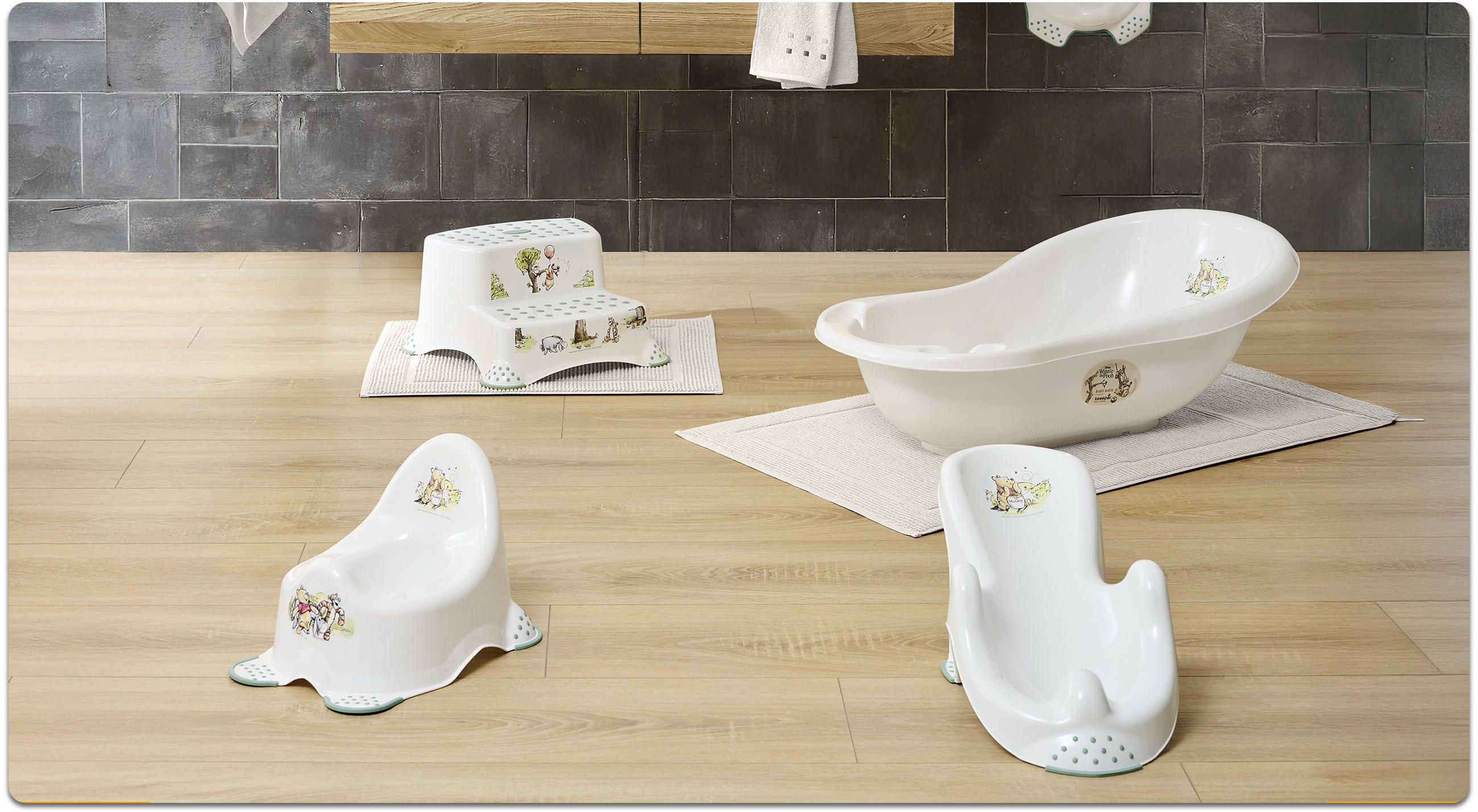 Weißes Baby-Badezubehör-Set mit kleinem Waschbecken, Töpfchen und Hocker auf Holzoberfläche vor grauen Fliesen, mit kleinen Tiermotiven.