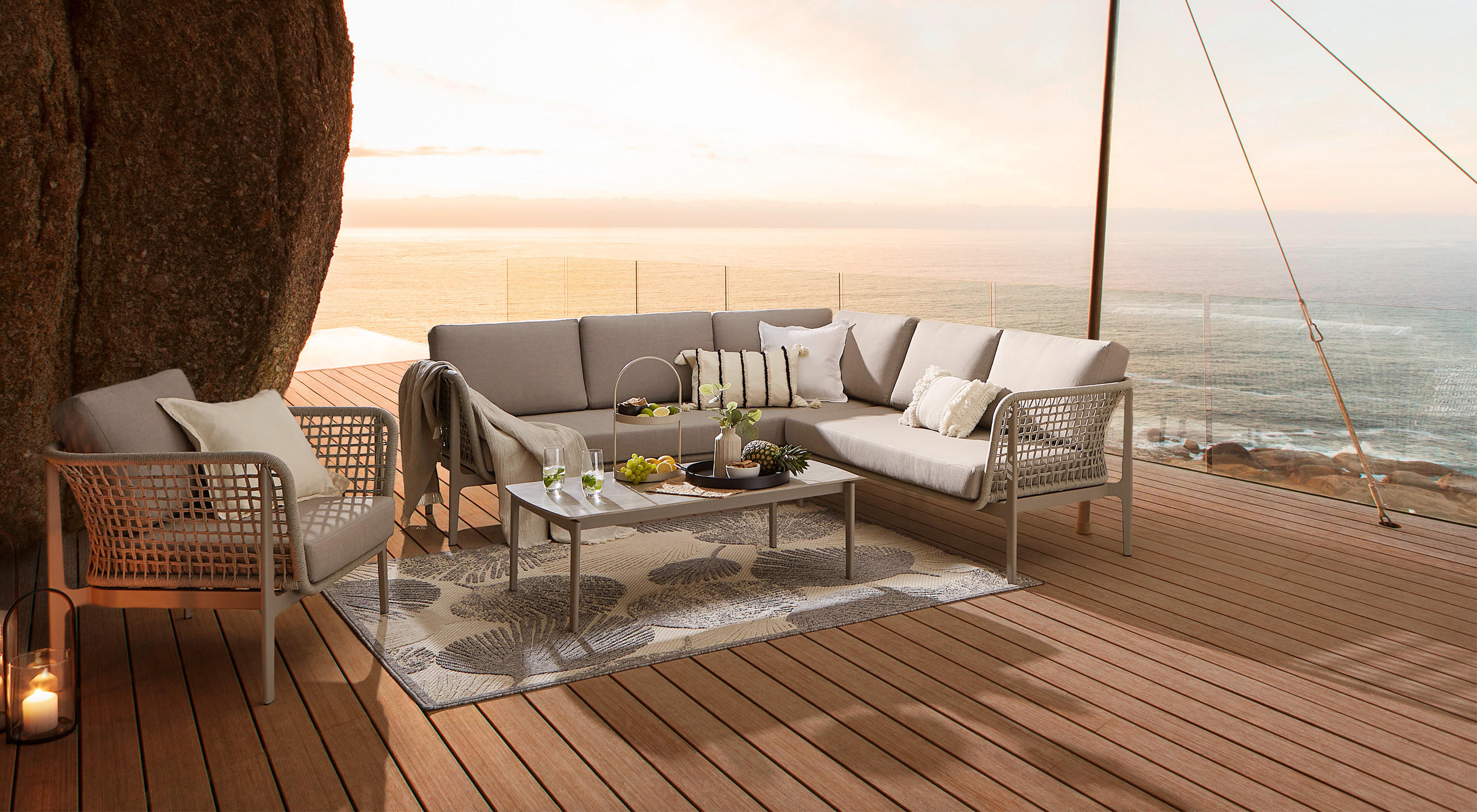 Terrasse mit Holzfußboden, Ecksofa mit Kissen, Sessel und Tisch mit Dekoration vor Meerblick bei Sonnenuntergang.