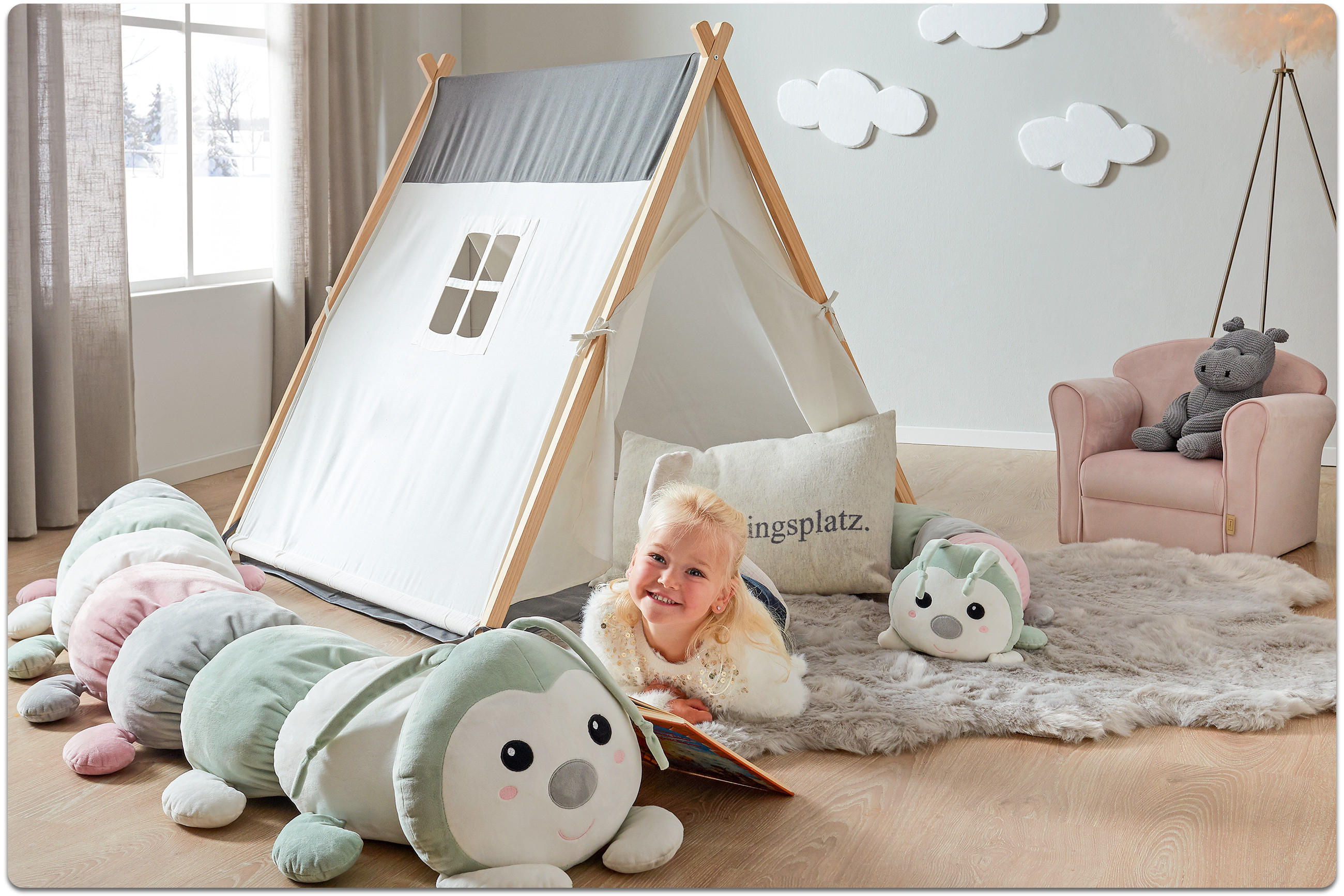 Kind liegt auf dem Boden vor einem Zeltspielhaus mit Fenster und liest ein Buch neben einem großen, weichen Raupenkuscheltier in einem hellen Kinderzimmer mit Teppich, Sessel und Wolkenwanddekoration.